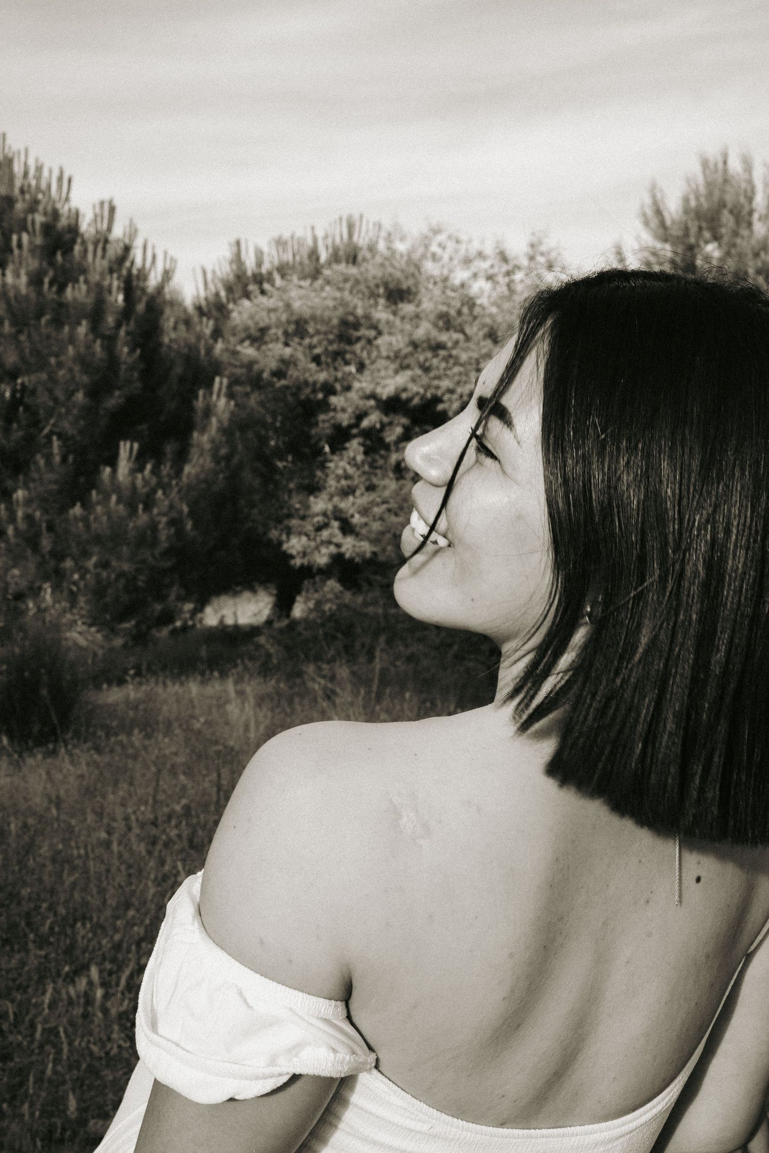 Black and white photo of a smiling woman with a bob haircut seen from behind, off-shoulder top, looking aside in a grassy field with trees.