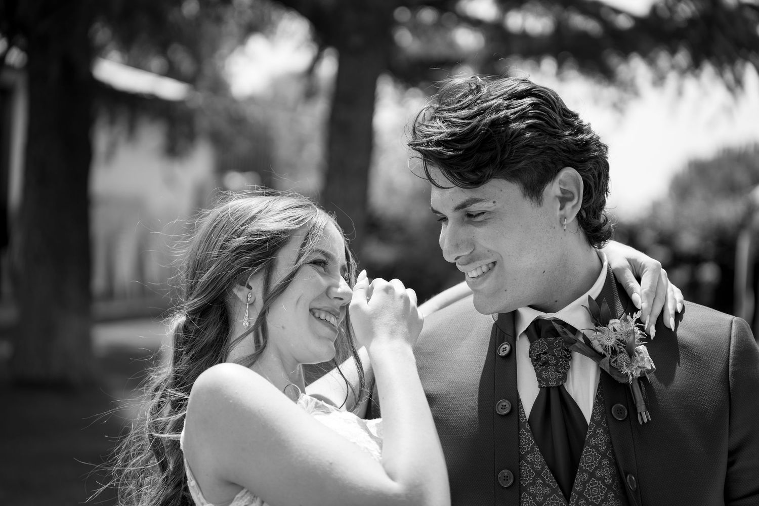 Black and white photo of a smiling young couple sharing a joyful moment at a wedding, the groom in a vest and floral boutonniere.