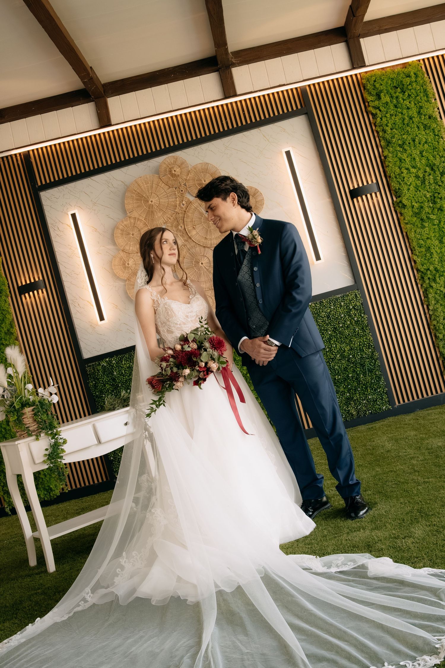 Bride in a white lace gown with a long veil holding a red bouquet beside a groom in a navy suit at an elegant garden wedding.
