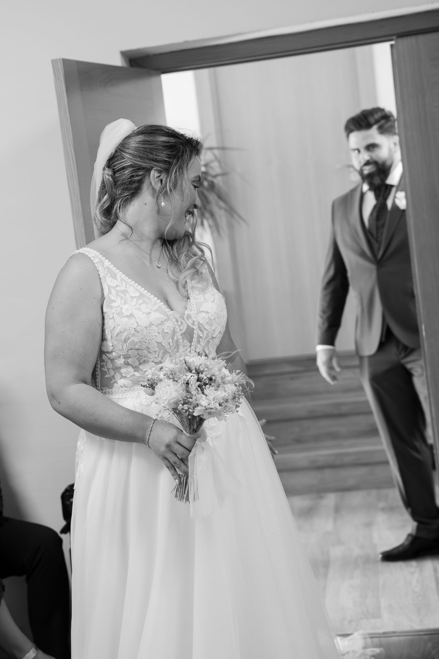 Black and white photo of a bride in a lace wedding dress holding a bouquet as the groom in a suit watches from a doorway.