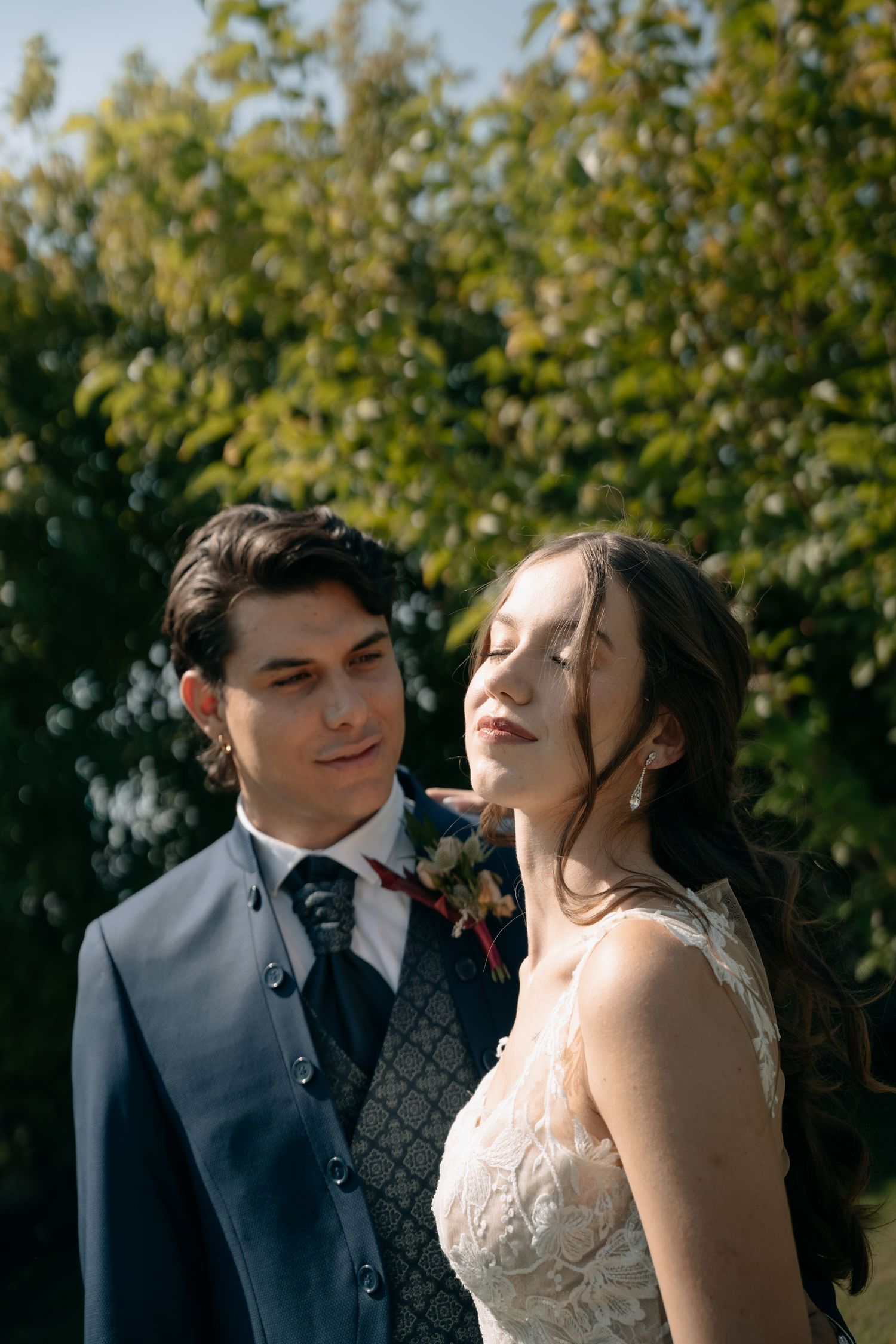 Groom in a navy suit with a floral boutonniere gazes at his bride in a lace wedding dress as she closes her eyes, with green trees behind.