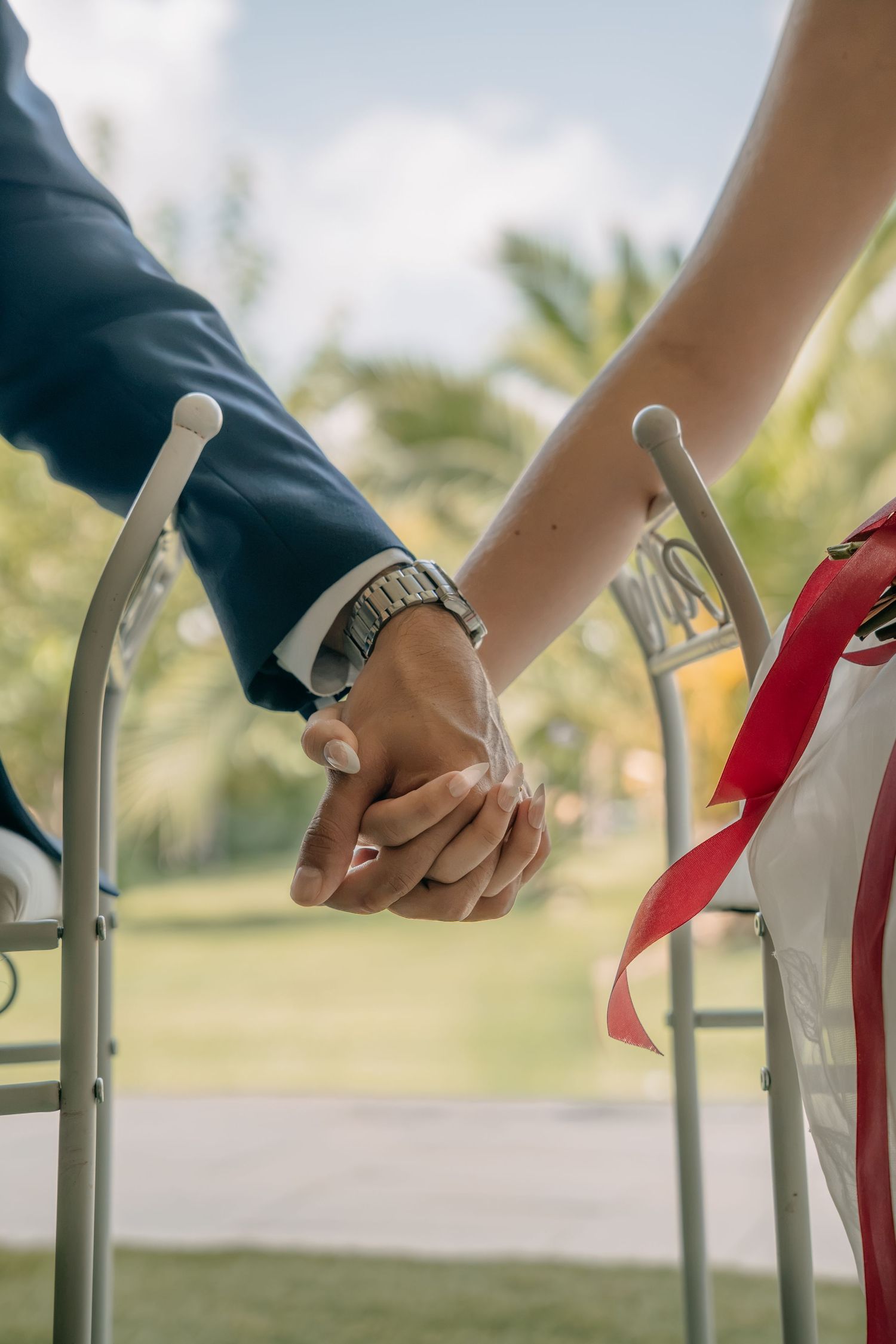 Couple holding hands between two chairs at an outdoor wedding ceremony, with a red ribbon and palm trees in the background.