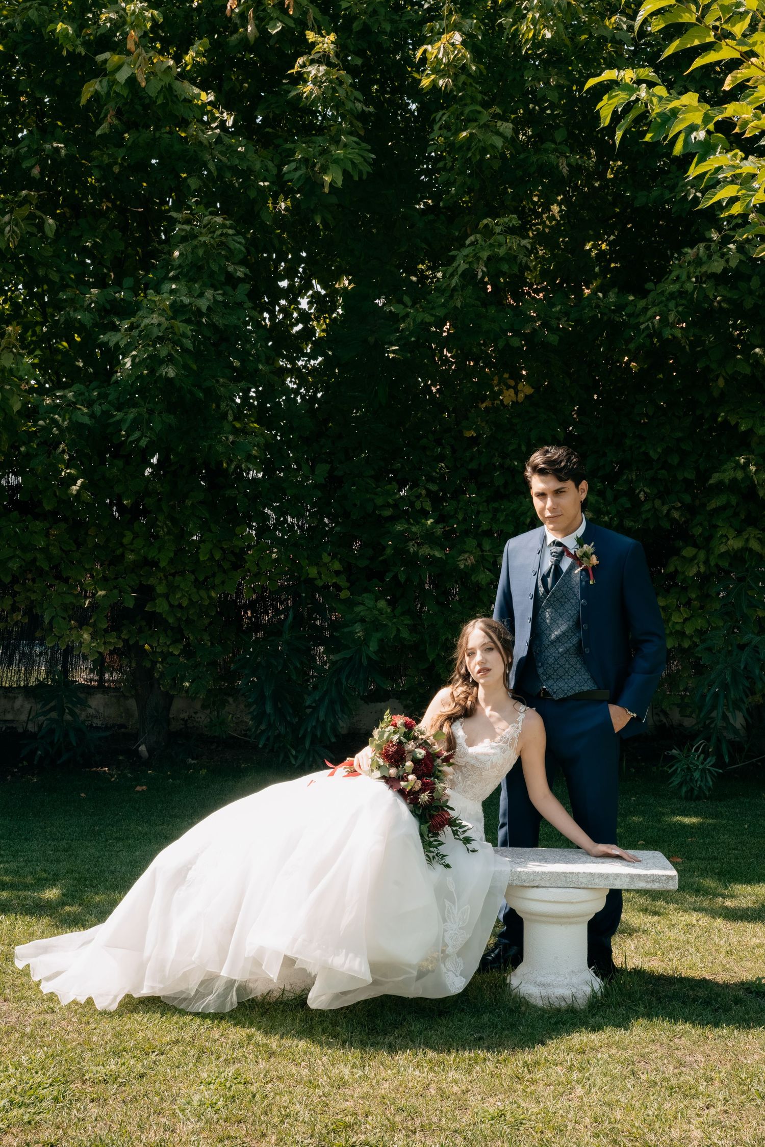 Bride in a flowing white gown holding a red bouquet reclines on a white bench beside a groom in a navy suit on a sunlit lawn.
