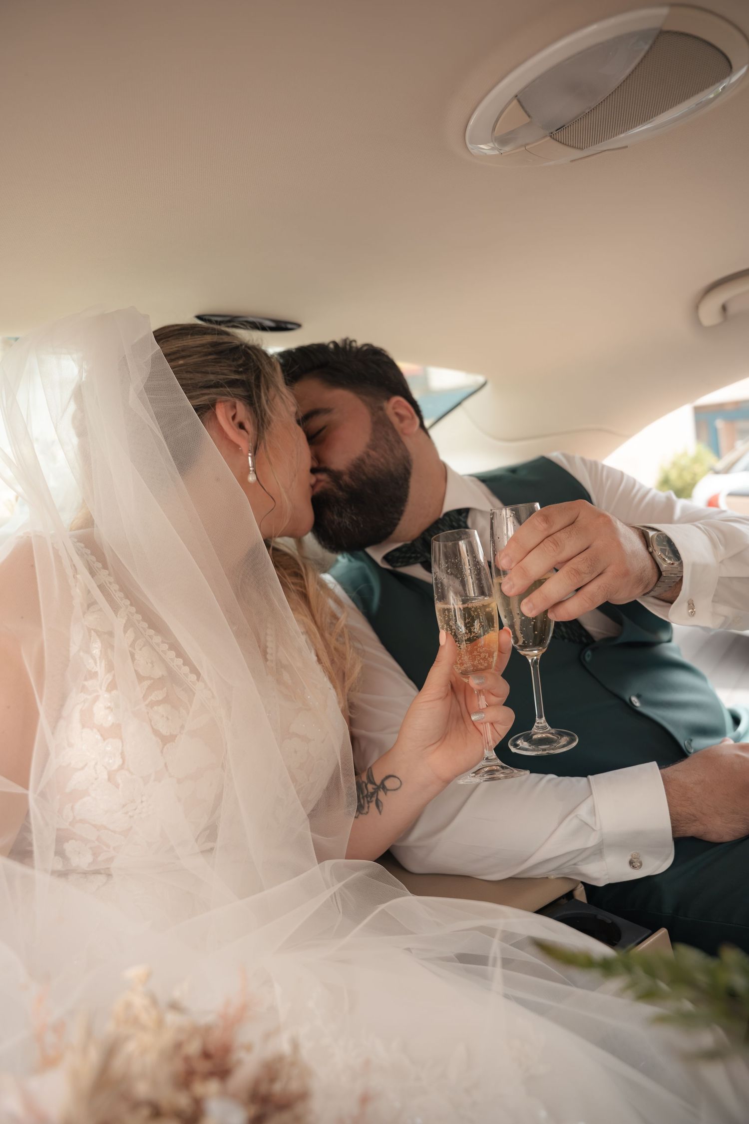 Bride in a lace gown and veil kisses her bearded groom in a green vest while they toast champagne glasses inside a car.