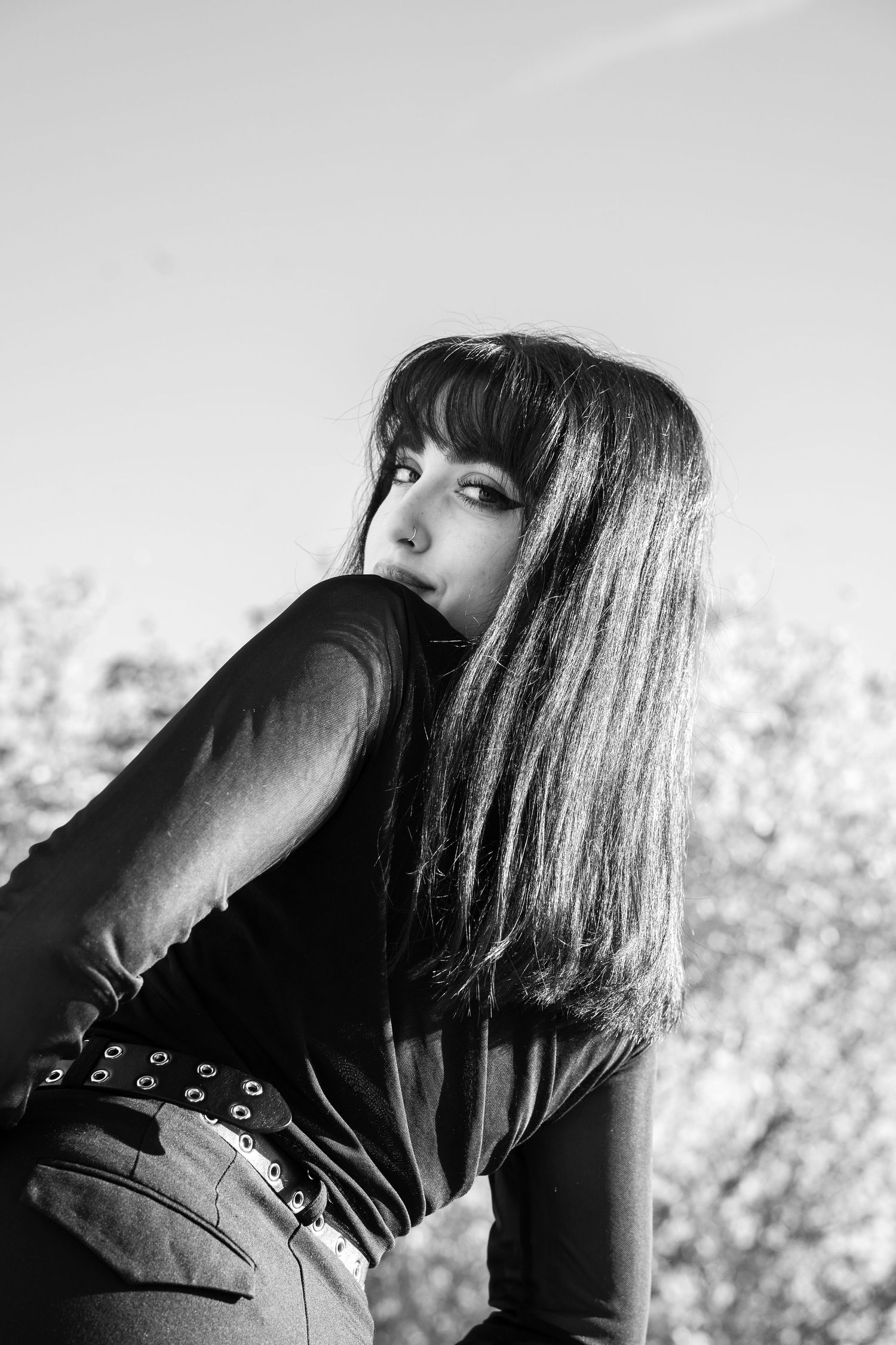 Black and white portrait of a woman with long dark hair and bangs glancing over her shoulder, wearing a studded belt against a bright sky.