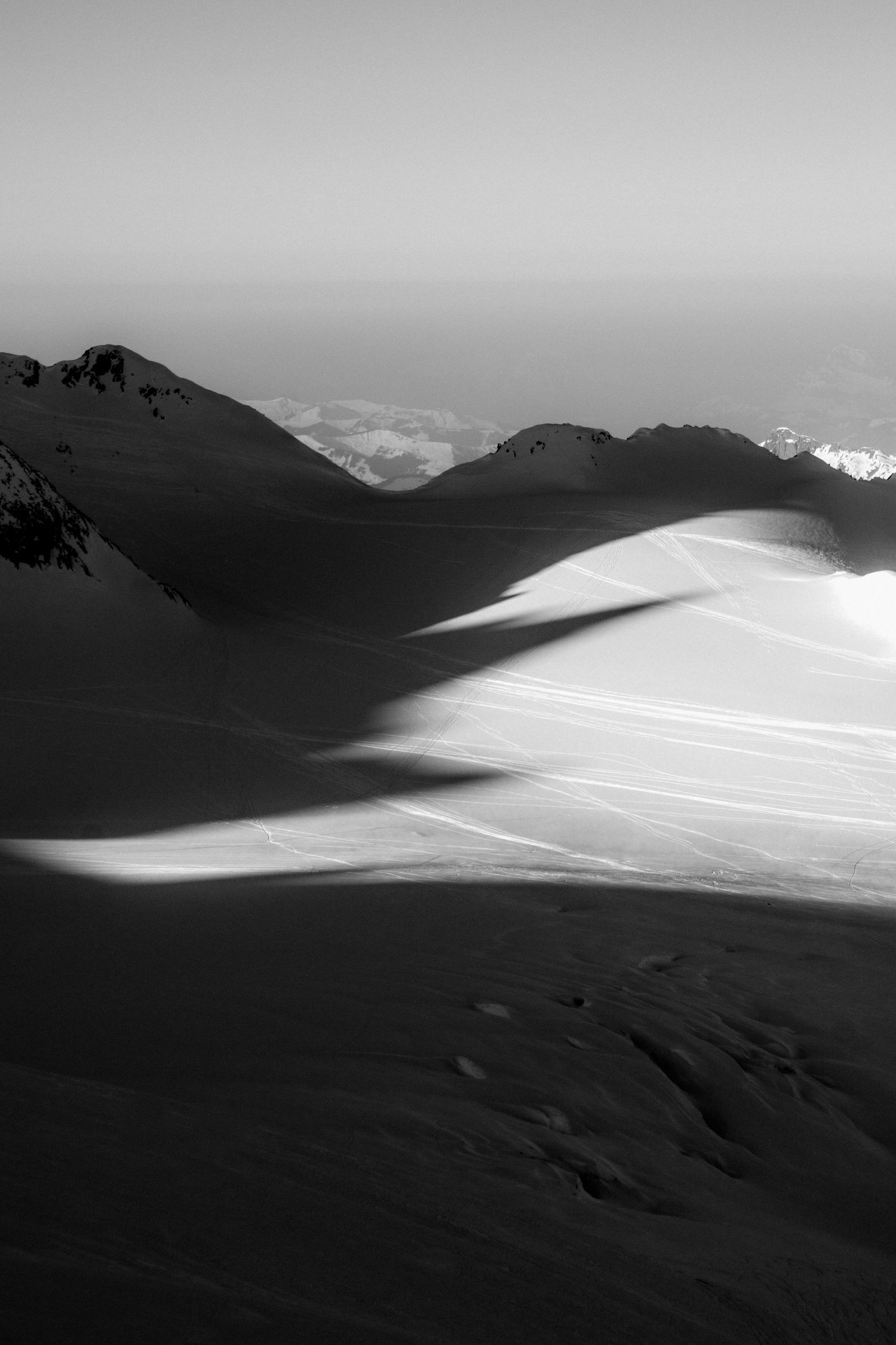 Black and white mountain landscape with dramatic shadows on snow-covered slopes and distant peaks under a clear sky.