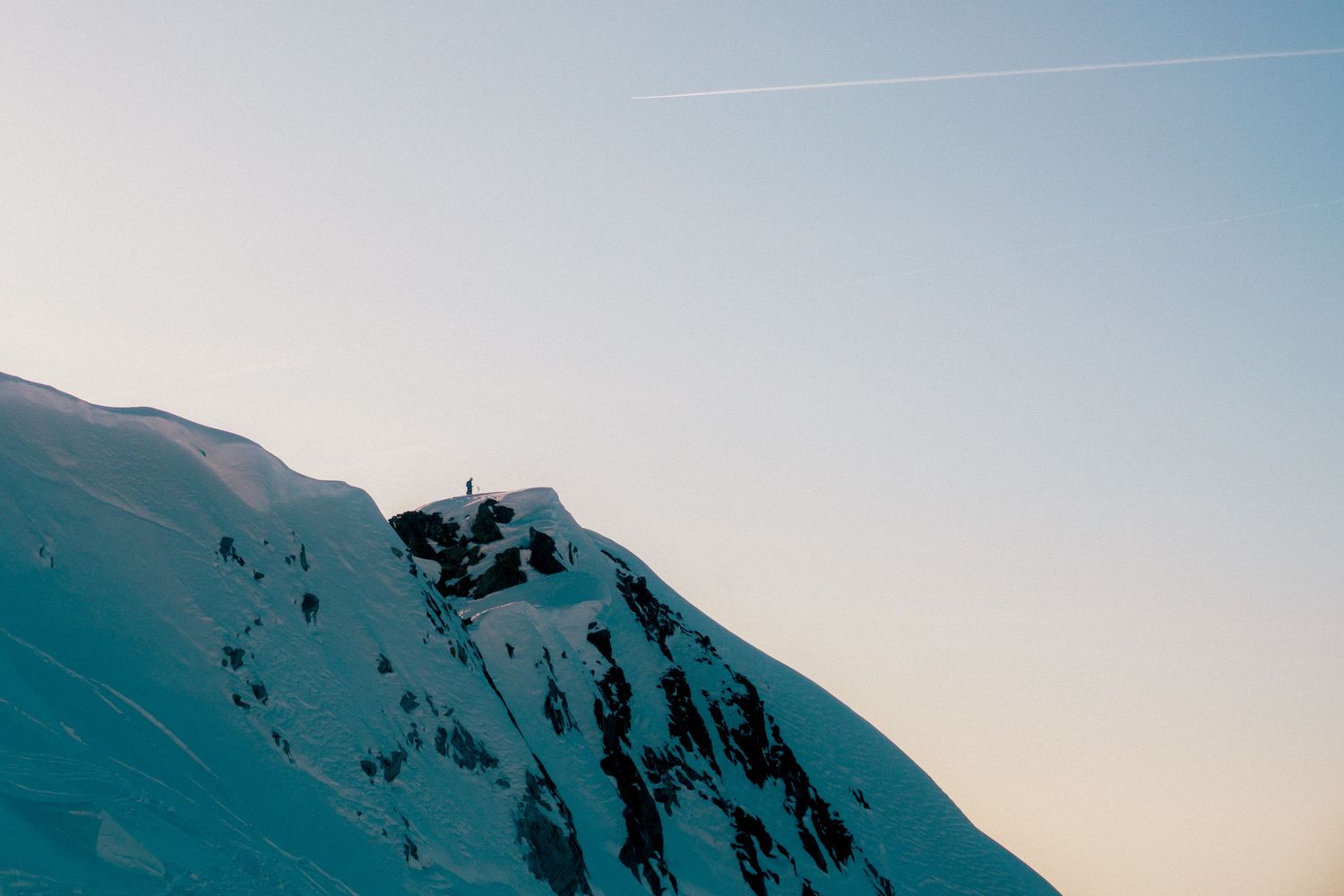 Snow-covered mountain peak at dusk with a lone figure standing on the summit against a pale sky.