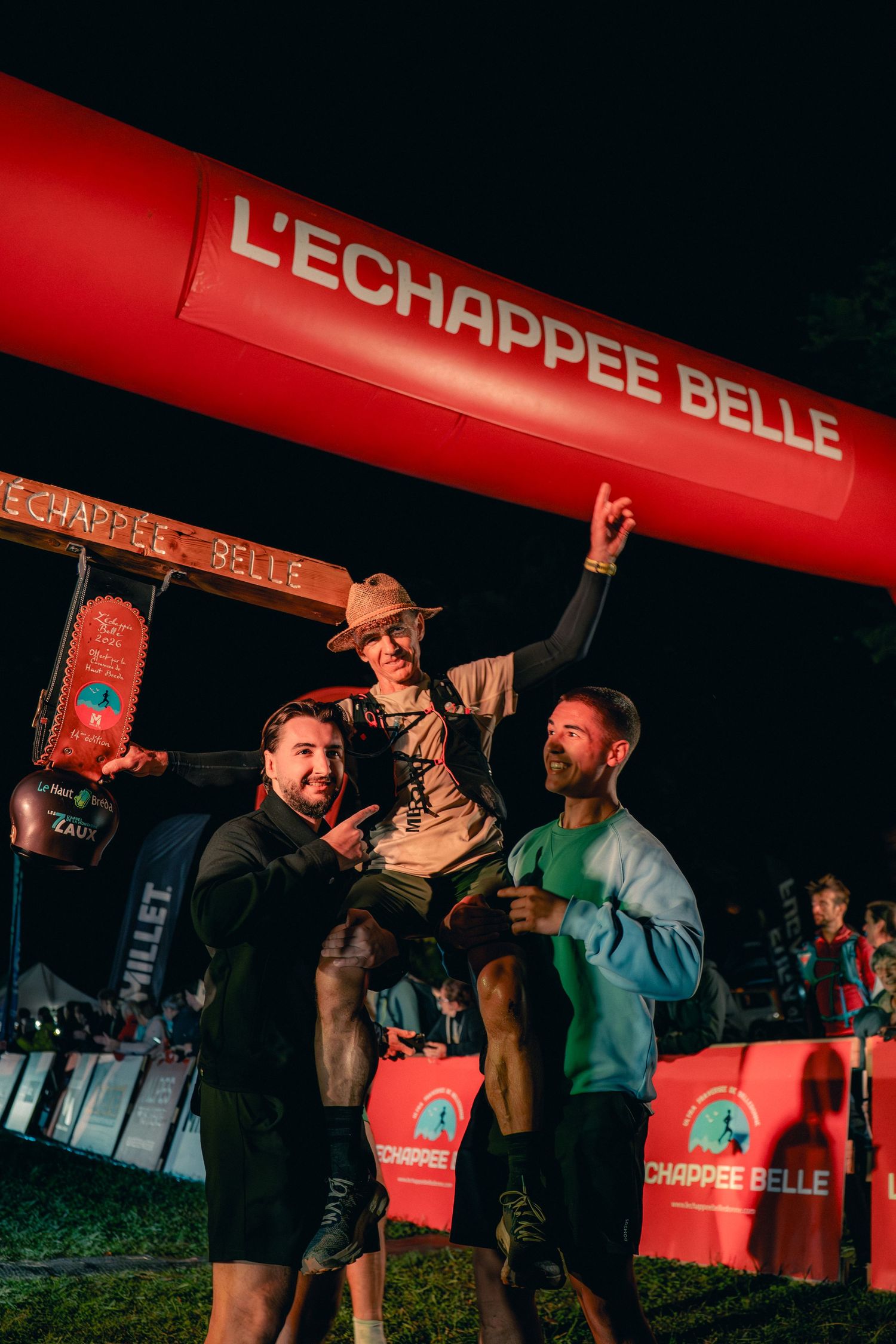 Victorious male trail runner lifted by two supporters under L'Echappee Belle race finish arch at night.