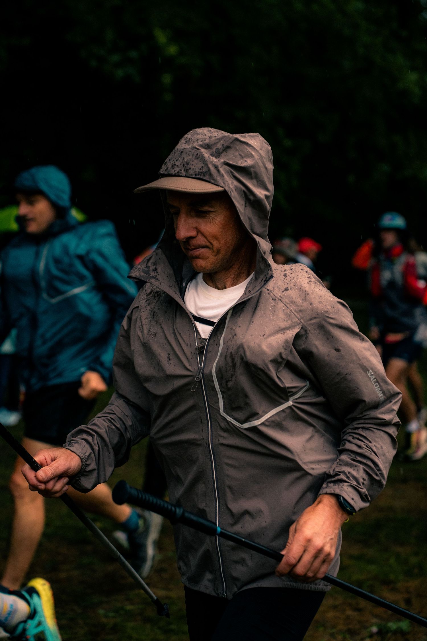 Muddy trail runner in gray hooded jacket holds trekking poles during rainy race.