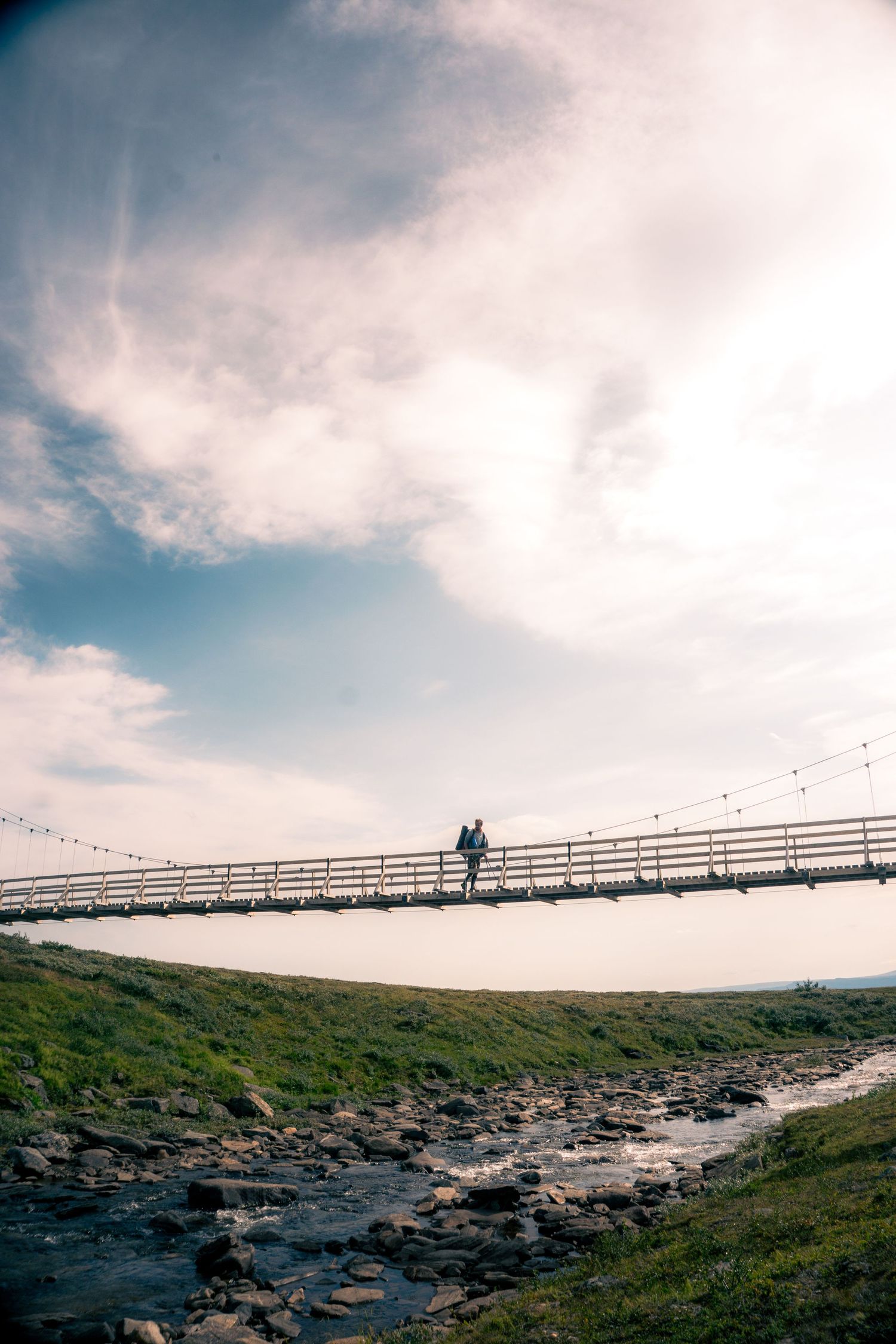 A hiker crosses a wooden suspension bridge over a rocky stream under a cloudy sky.