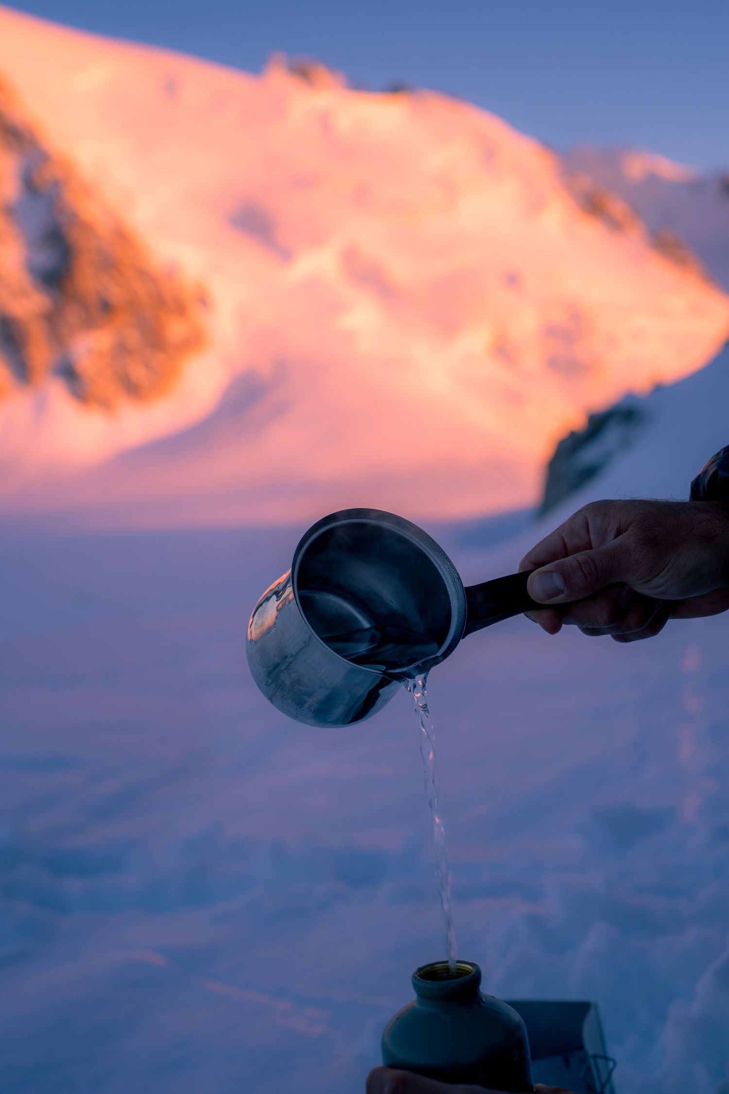 Hand pouring water from a metal pot outdoors in snow with a pink alpenglow mountain in the background.