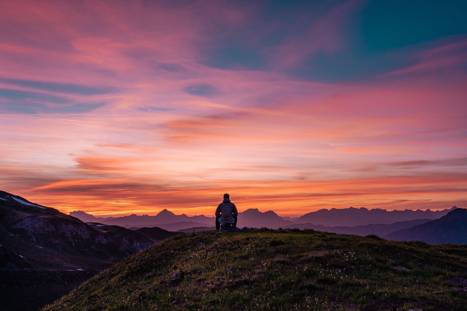 Solitary figure sits on mountain hilltop watching vibrant pink and orange sunset over distant peaks.