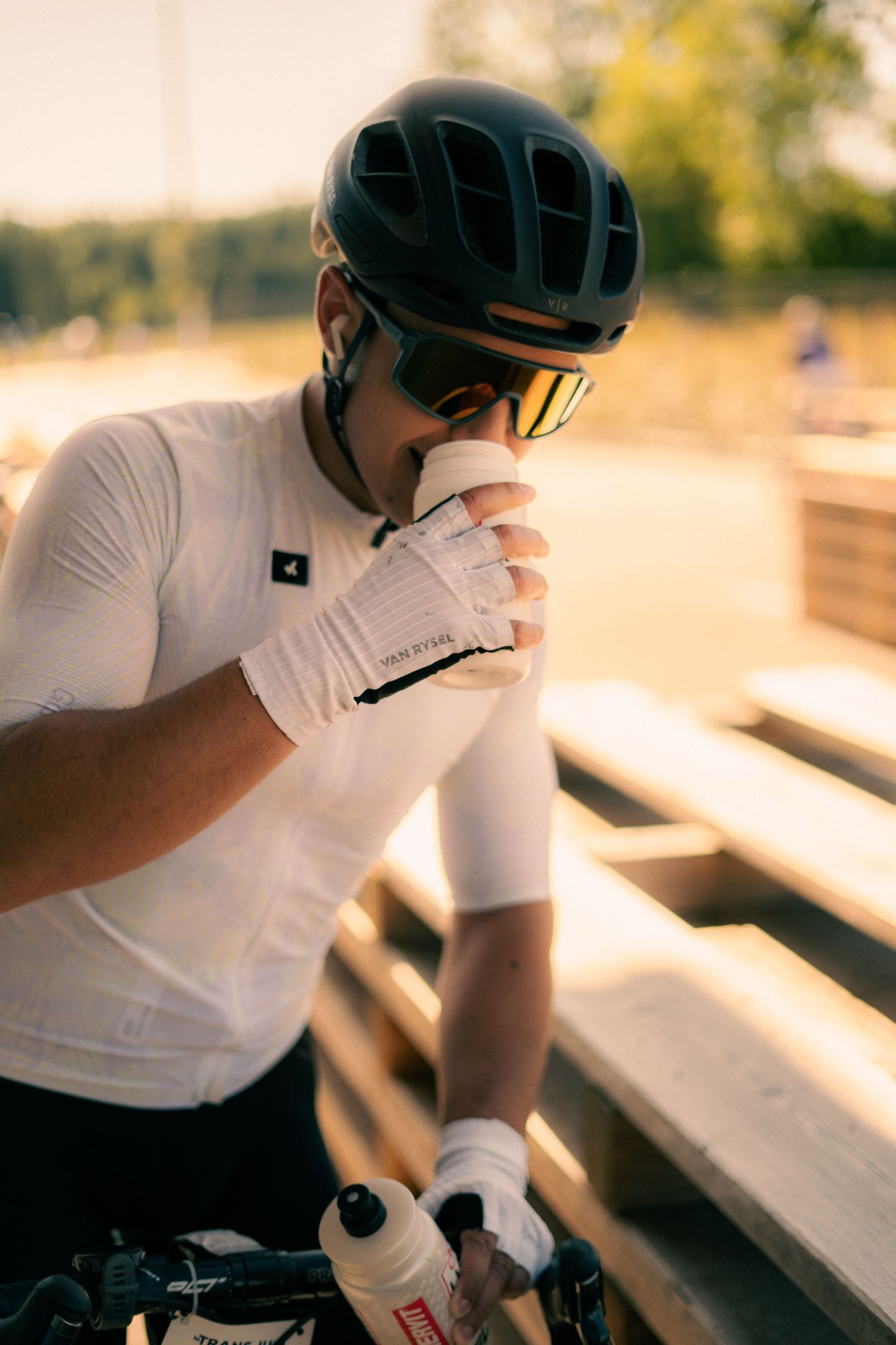 Cyclist in white jersey and black helmet drinks from cup while holding water bottles outdoors.