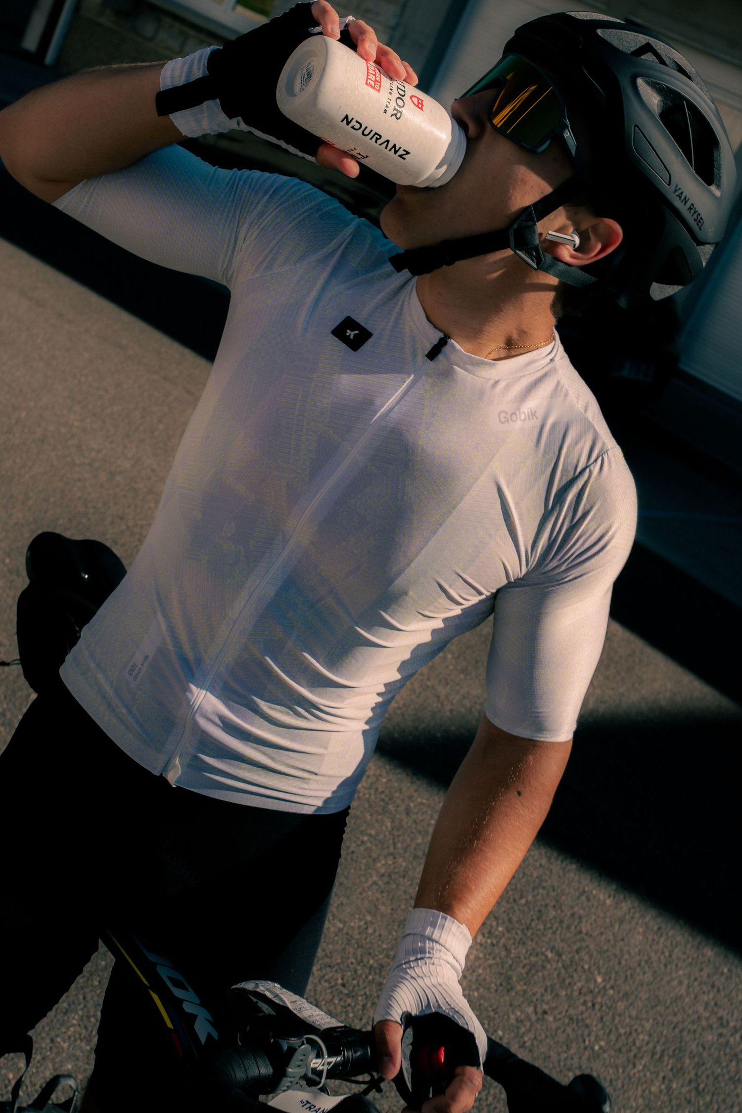 Cyclist in white jersey and helmet drinking from a water bottle during a rest stop outdoors.