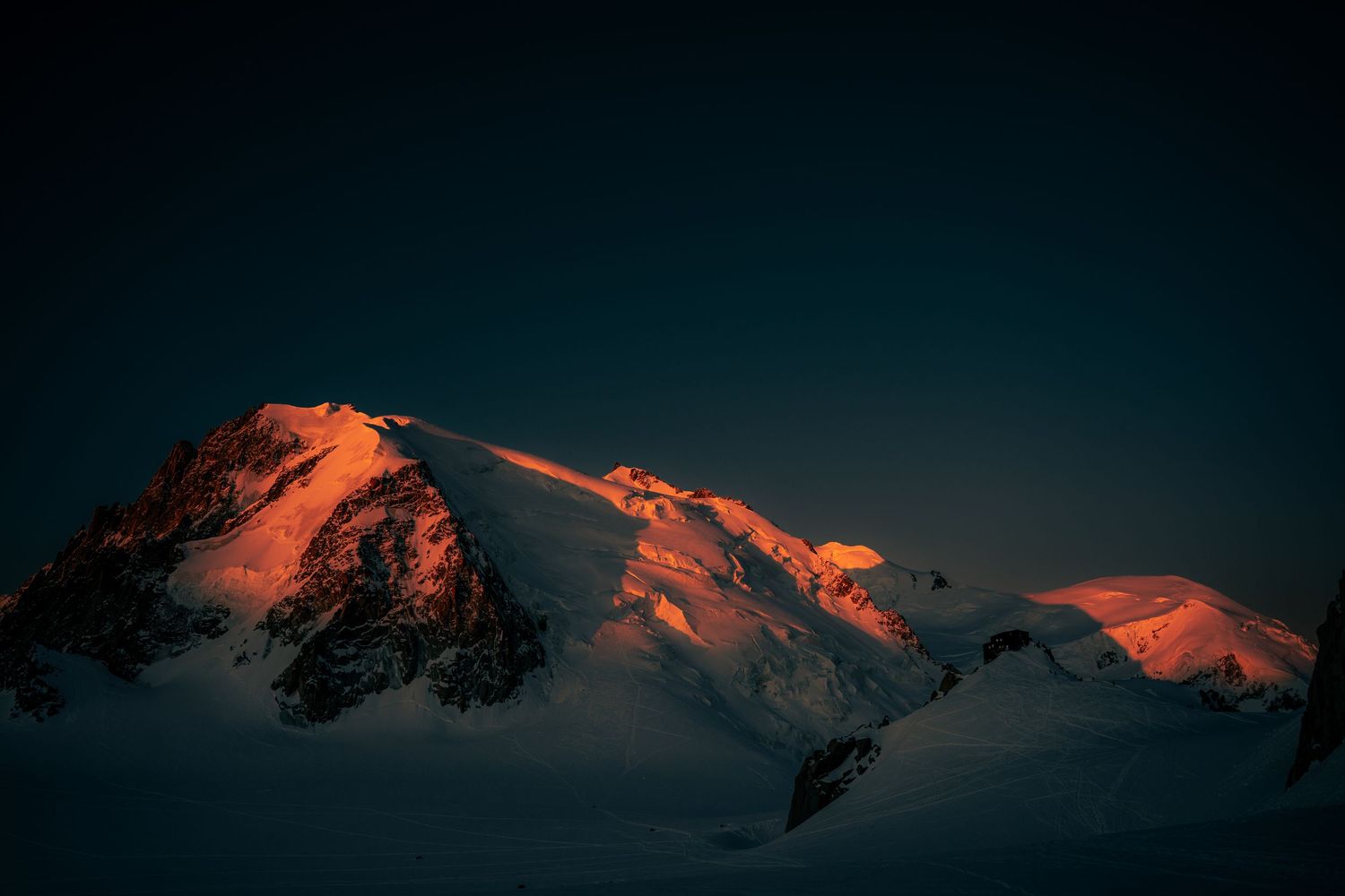 Twin snow-covered mountain summits glowing vivid orange in alpenglow against a deep dark sky.