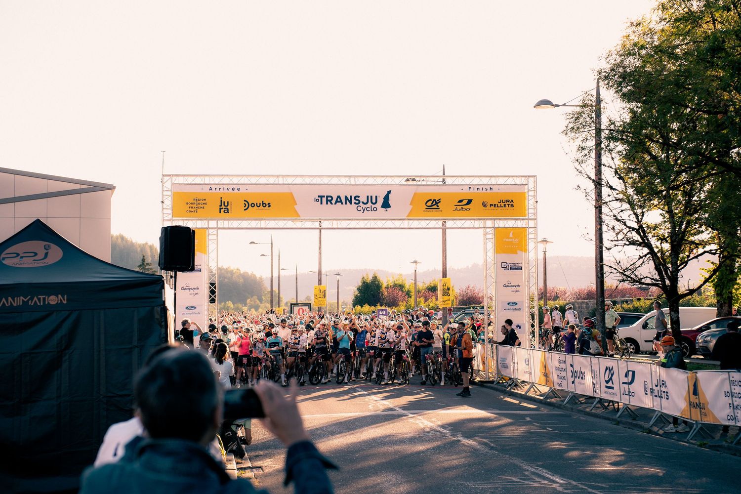Cyclists gather at the TransJu race start line under a banner, with spectators and a photographer in the foreground.