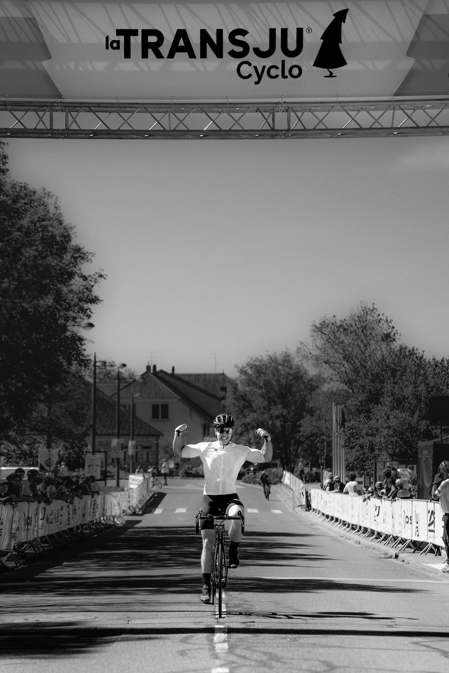 Cyclist celebrates victory crossing finish line at La TransJu Cyclo race, arms raised in triumph, black and white photo.