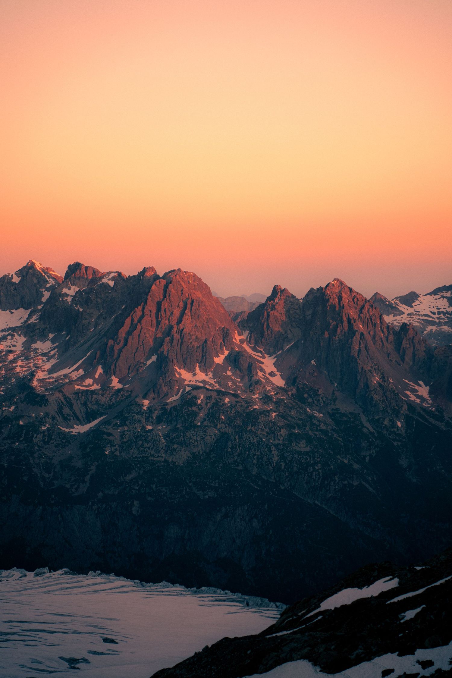 Jagged snow-covered mountain peaks bathed in warm orange alpenglow light at sunset in alpine wilderness.