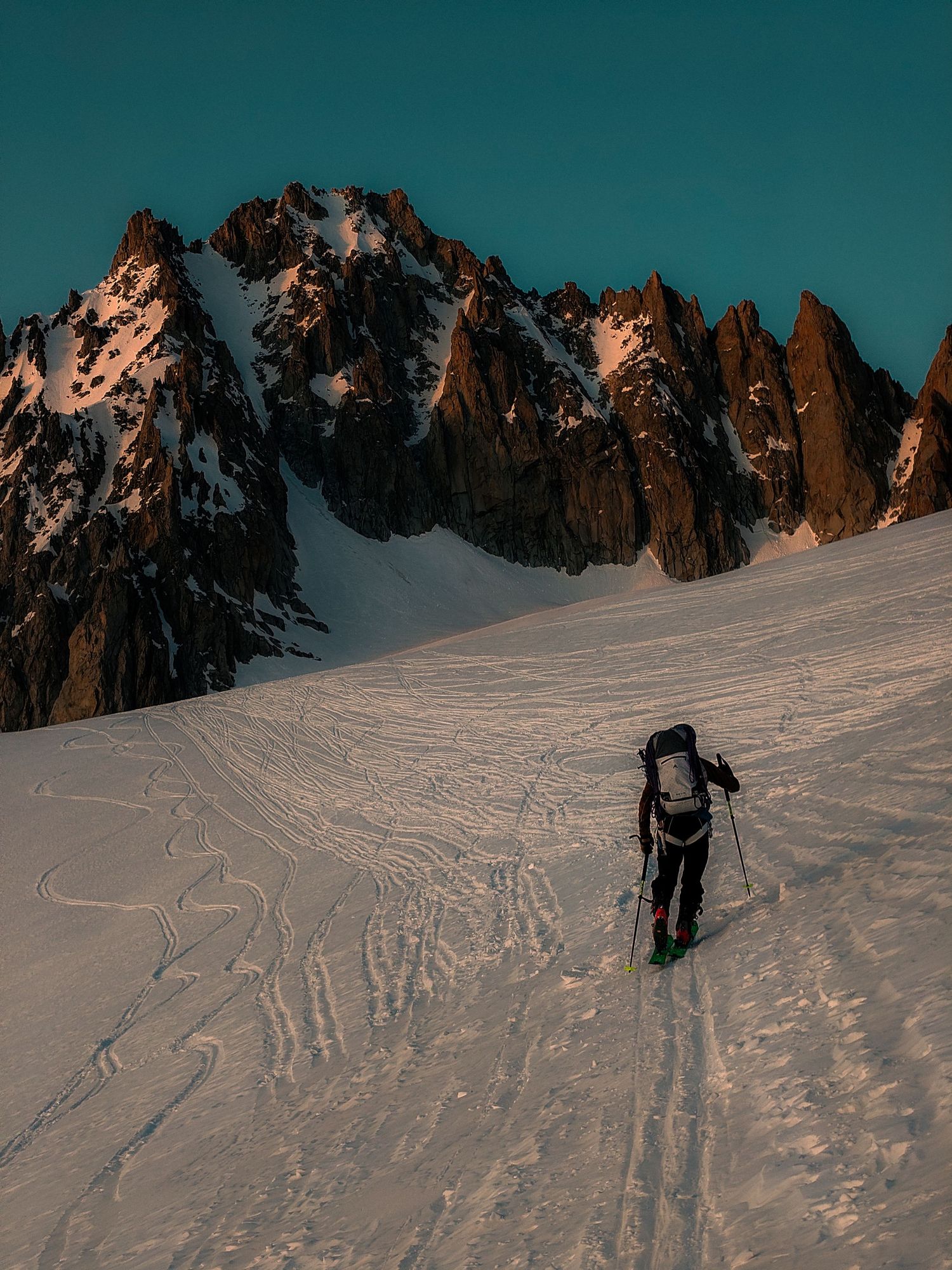 A lone skier ascends a snowy alpine slope with dramatic jagged mountain peaks in the golden hour light.