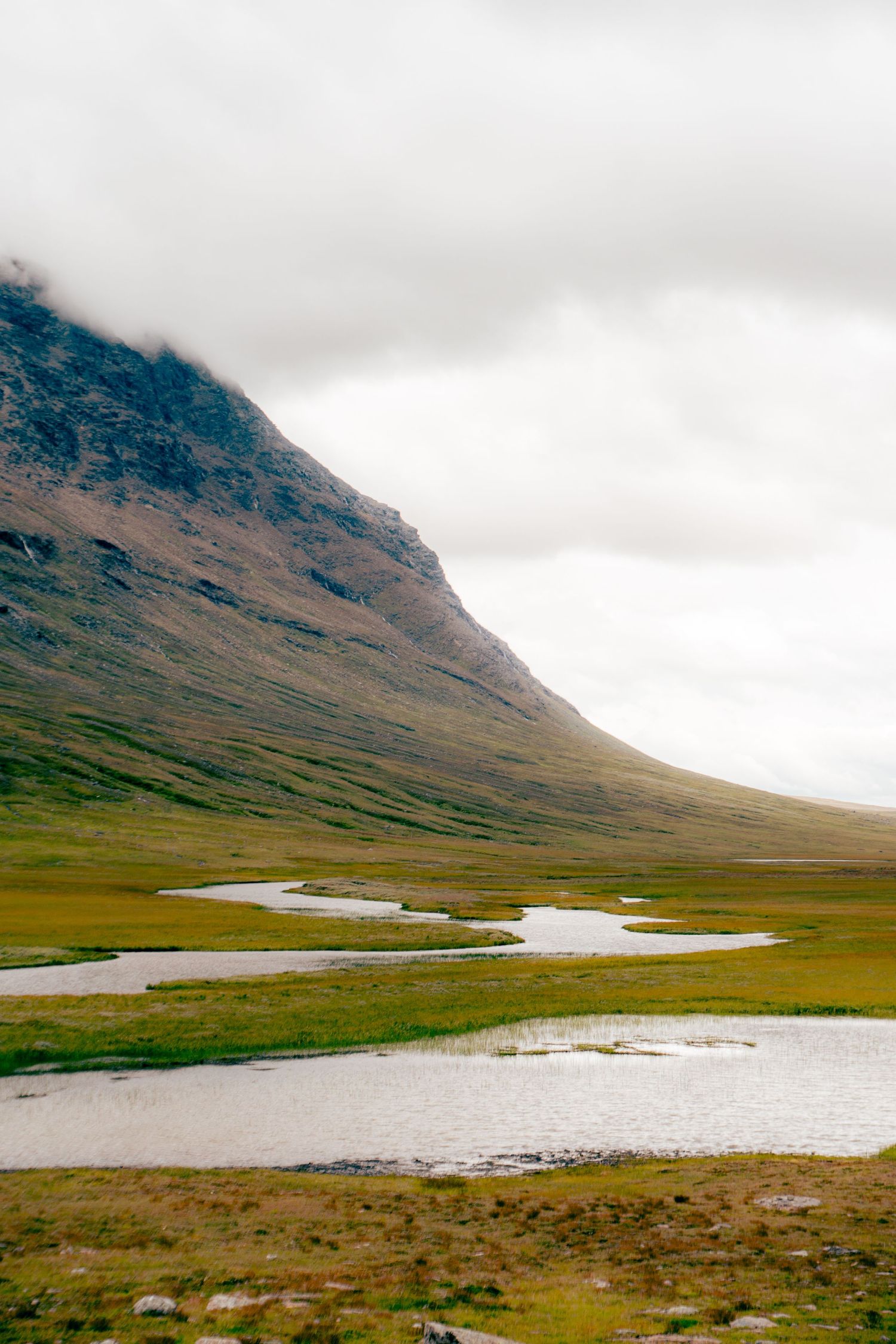 A winding river reflects the sky beside a steep mountain slope under cloudy skies.