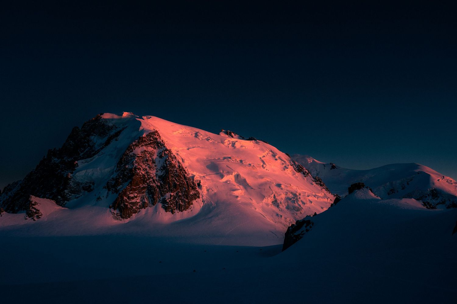 Snow-covered mountain summit illuminated by dramatic red alpenglow against a dark twilight sky.