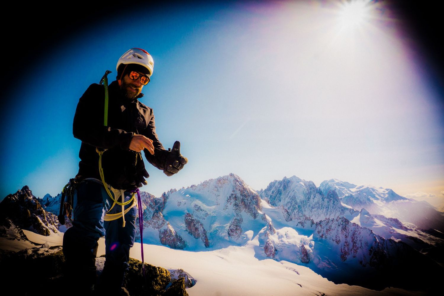 Mountain climber in helmet and gear stands on snowy alpine peak with dramatic sunlit mountain range behind.