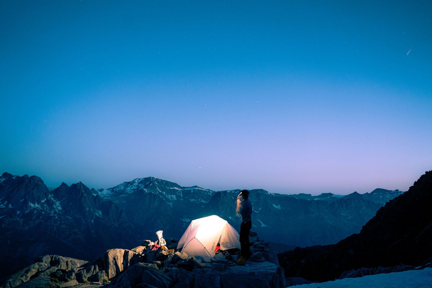 Lone camper stands beside illuminated tent on snowy mountain ridge under deep blue twilight sky.