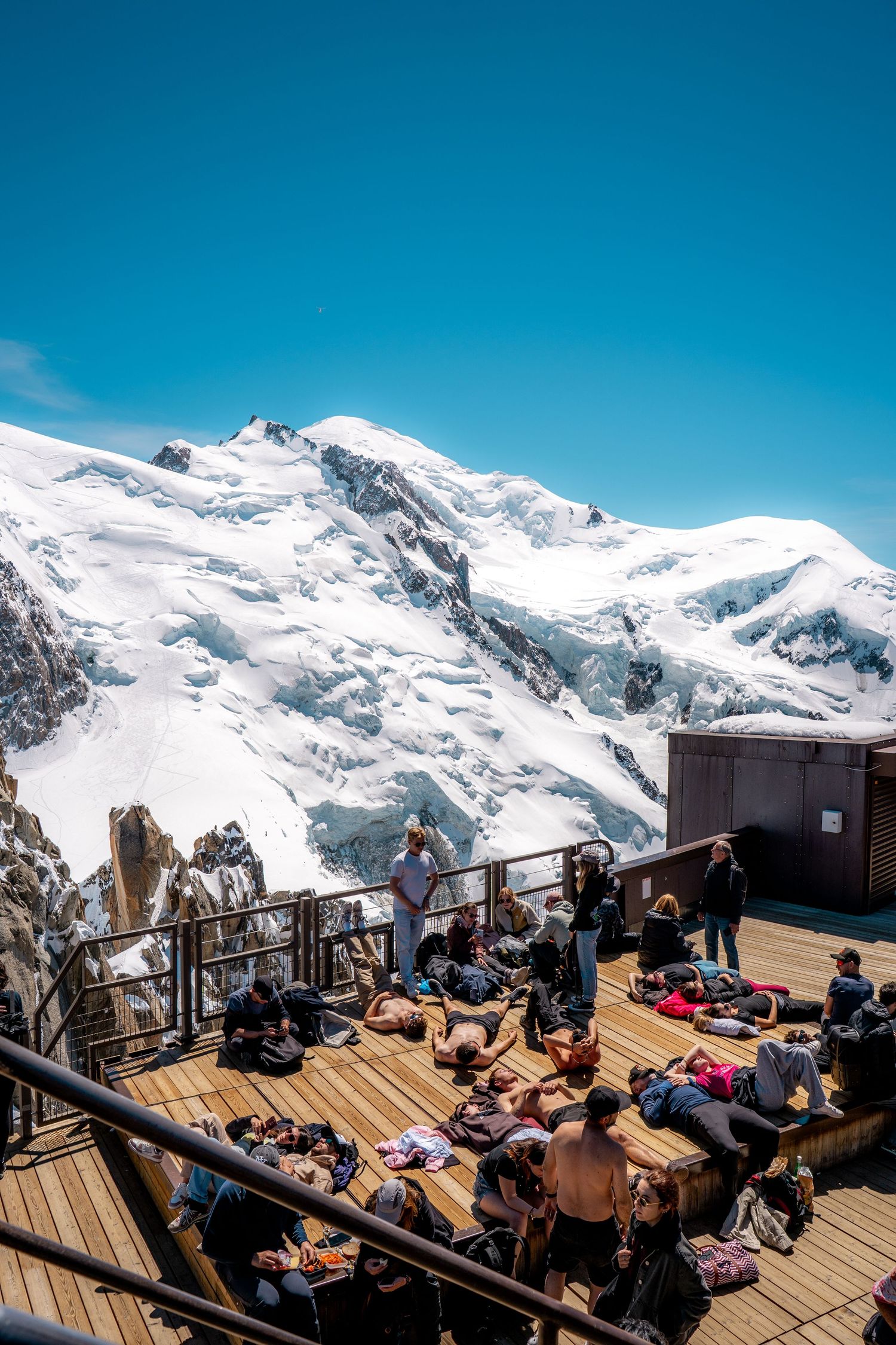 Tourists relaxing on sunny Aiguille du Midi terrace deck with massive Mont Blanc glacier backdrop.