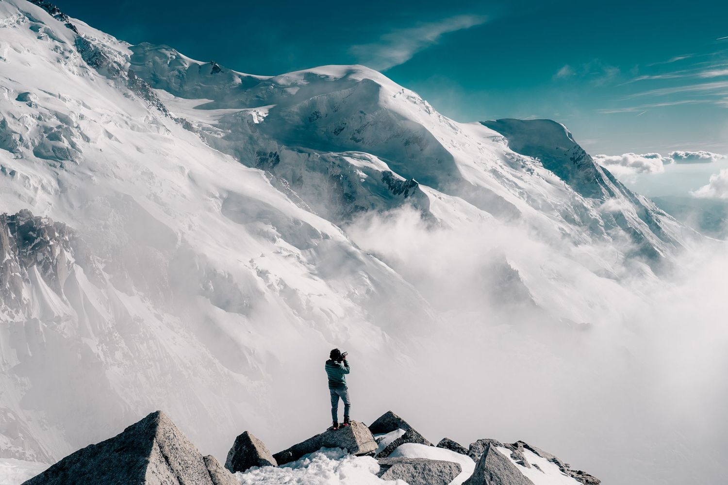 A solitary hiker stands on rocky outcrops overlooking dramatic snow-covered Mont Blanc peaks above the clouds.