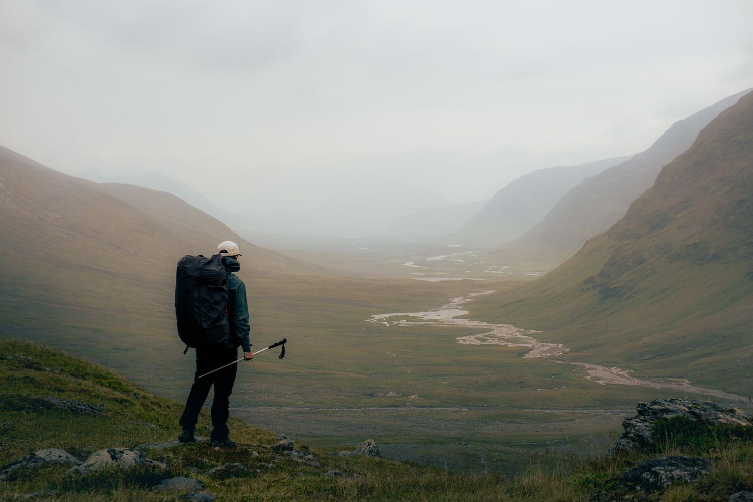 Solo hiker with backpack overlooking a misty valley with a winding river in remote Scottish Highlands.