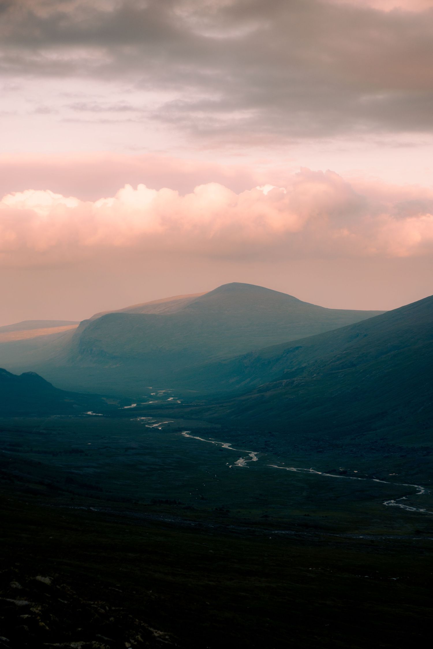 Serene mountain valley at sunset with a winding river and pink-hued clouds over distant peaks.