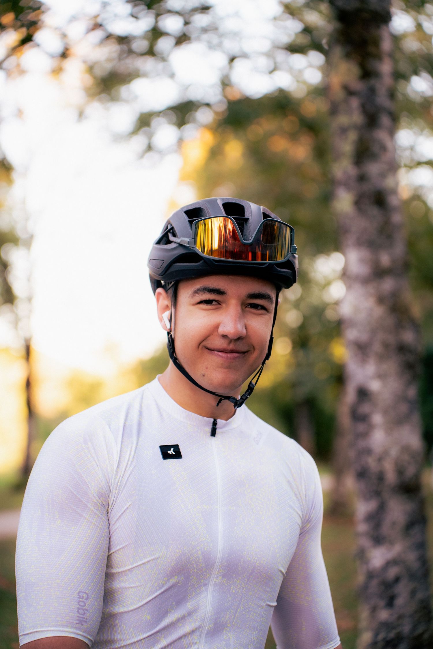 Young male cyclist smiling in a white jersey and black helmet with gold reflective goggles outdoors.