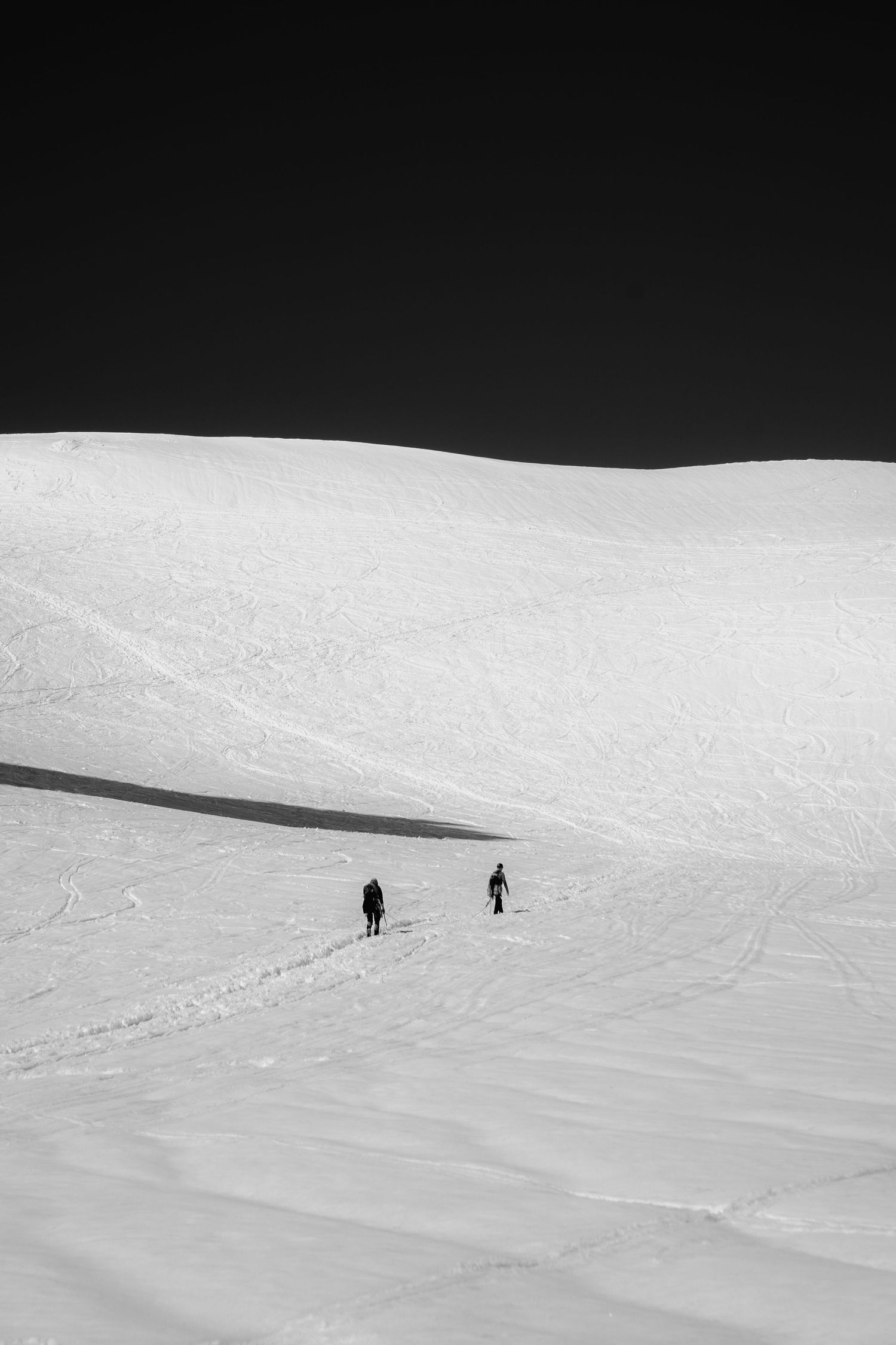 Two skiers traverse a vast snowy mountain slope in a minimalist black and white winter landscape.