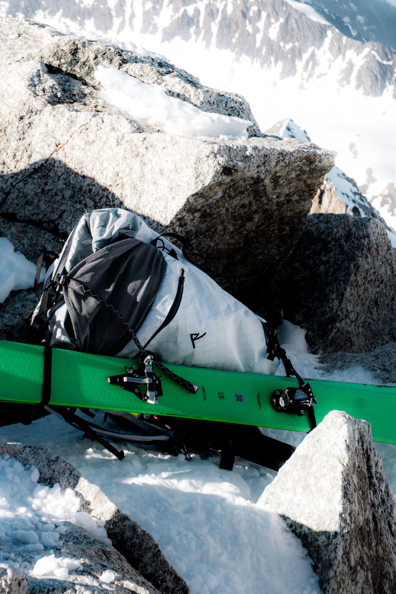 Green skis with bindings and a gray backpack resting on rocky snow-covered mountain terrain.