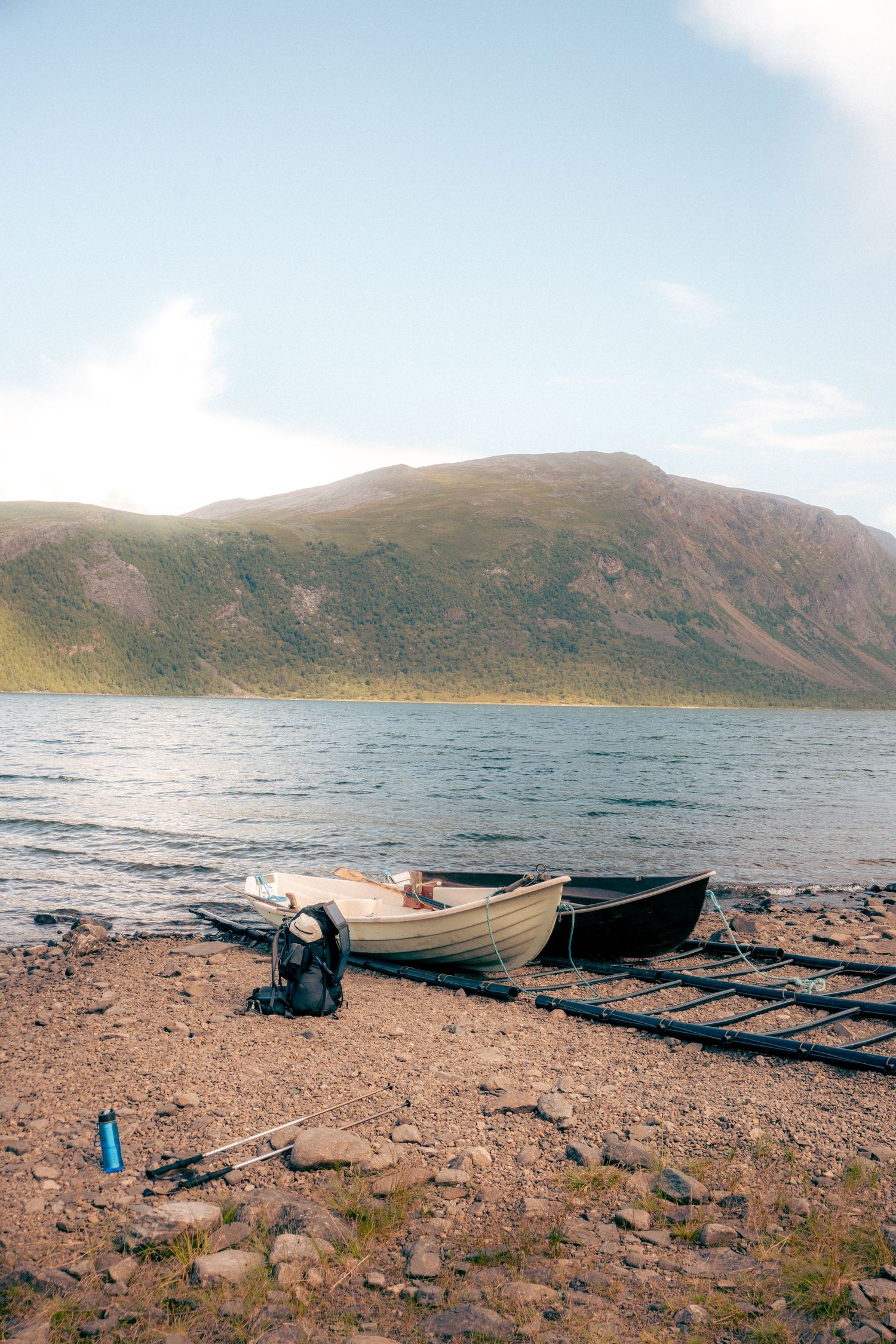 Two boats moored on a rocky lakeshore with a backpack and fishing rod beside a mountain.