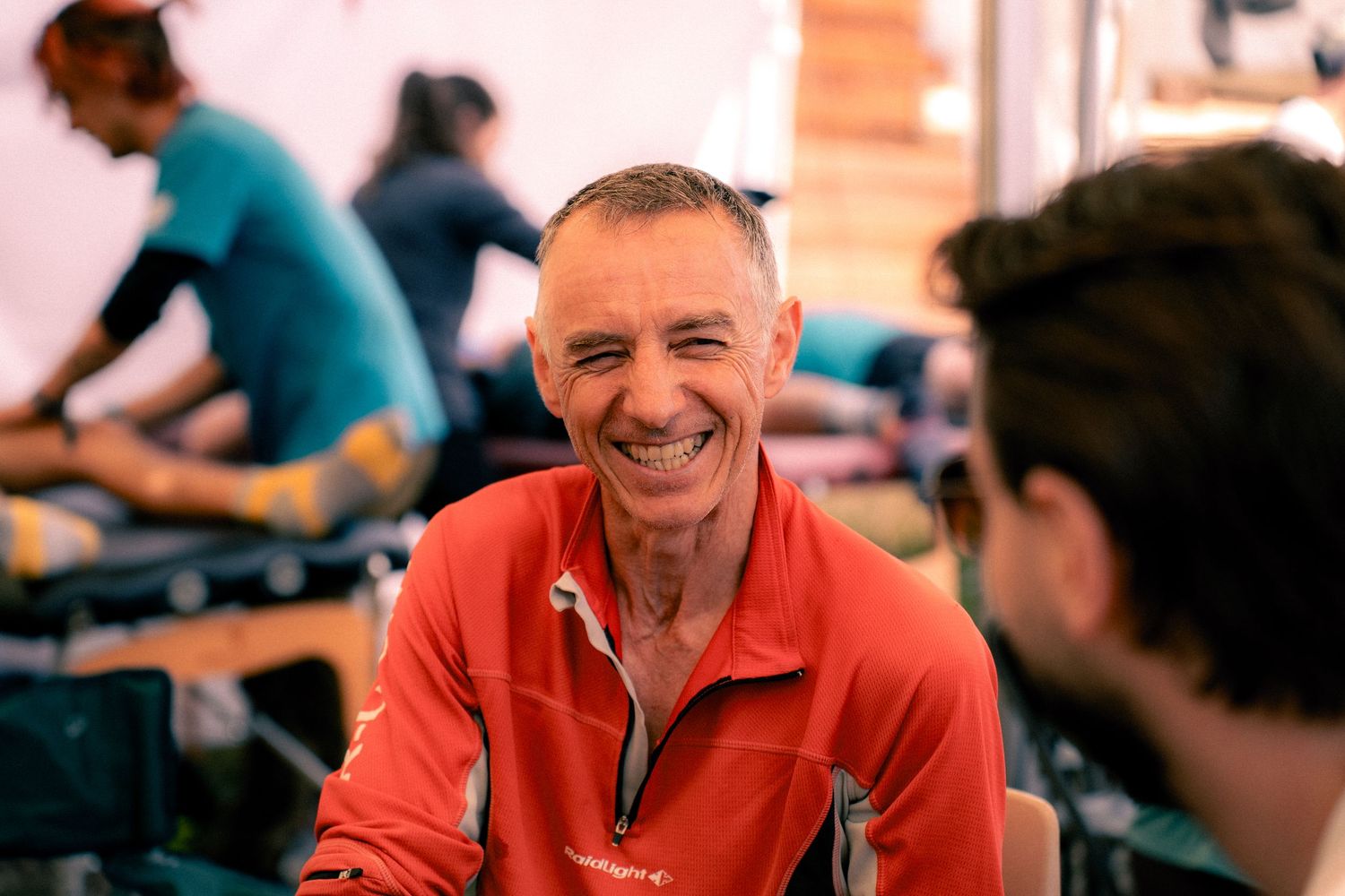Smiling male trail runner in red jacket relaxing at a race aid station during an ultramarathon event.