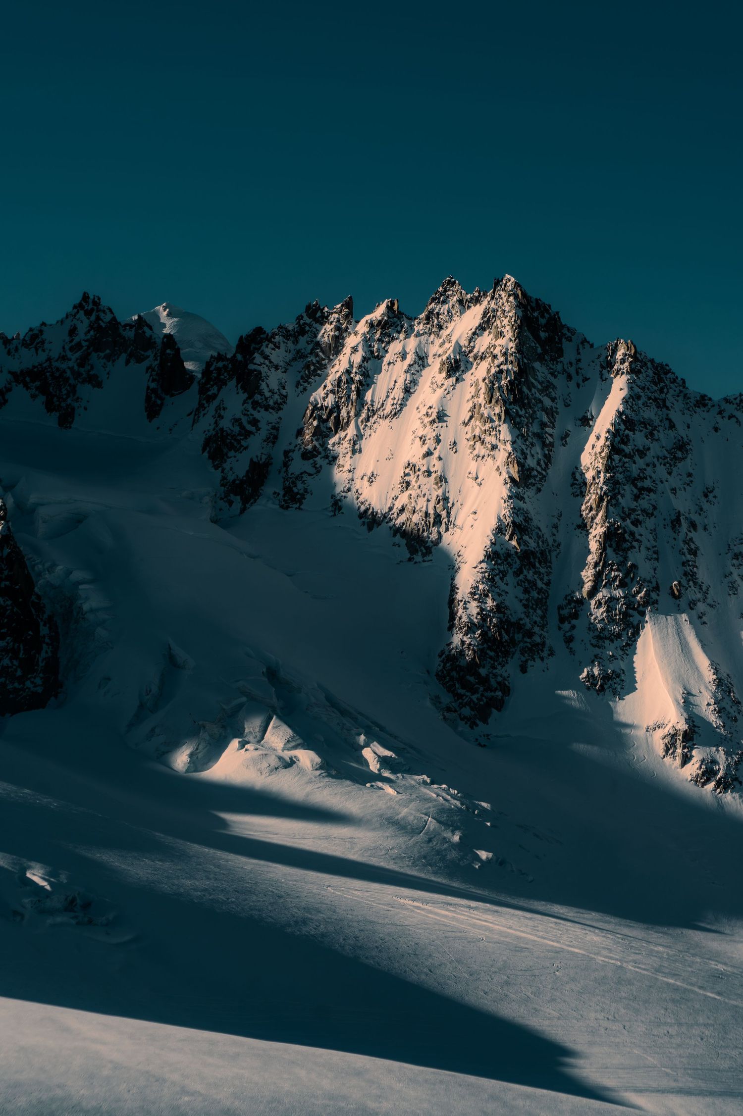 Snow-covered mountain peaks bathed in warm golden light against a clear blue sky in a dramatic alpine landscape.