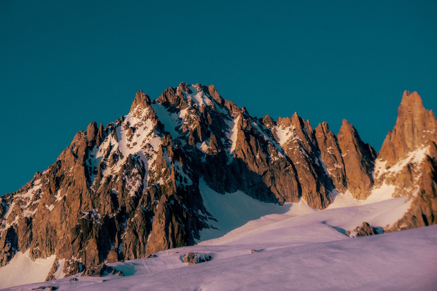 Snow-dusted rocky mountain peaks rise dramatically against a clear teal sky in a stunning alpine landscape.
