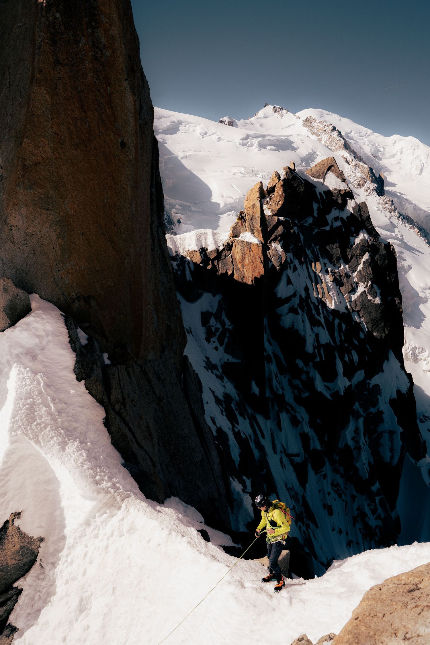 A mountaineer in a yellow jacket climbs a snowy ridge with rocky peaks and Mont Blanc in the background.