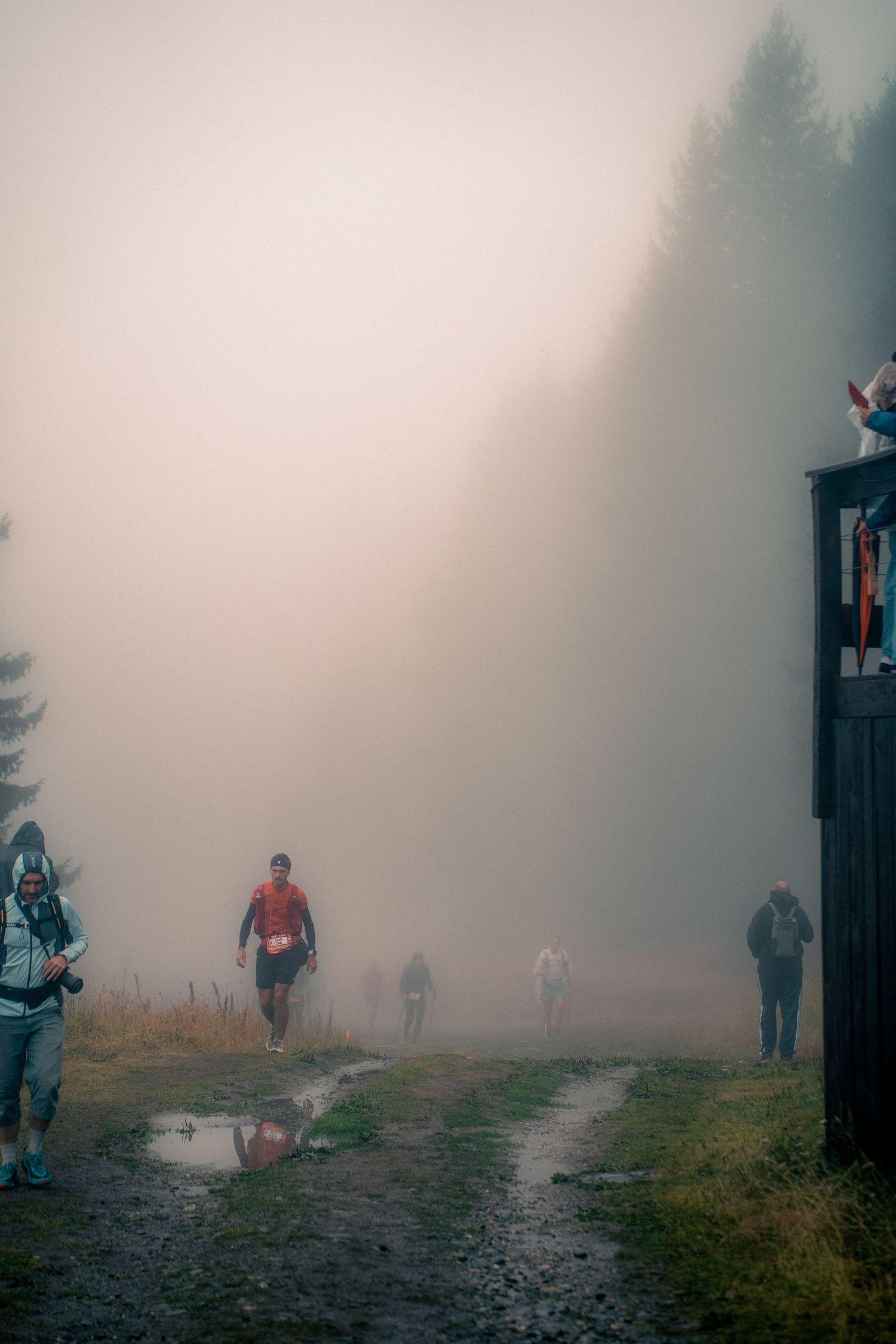 Trail runners emerge from dense fog on muddy mountain path during race.