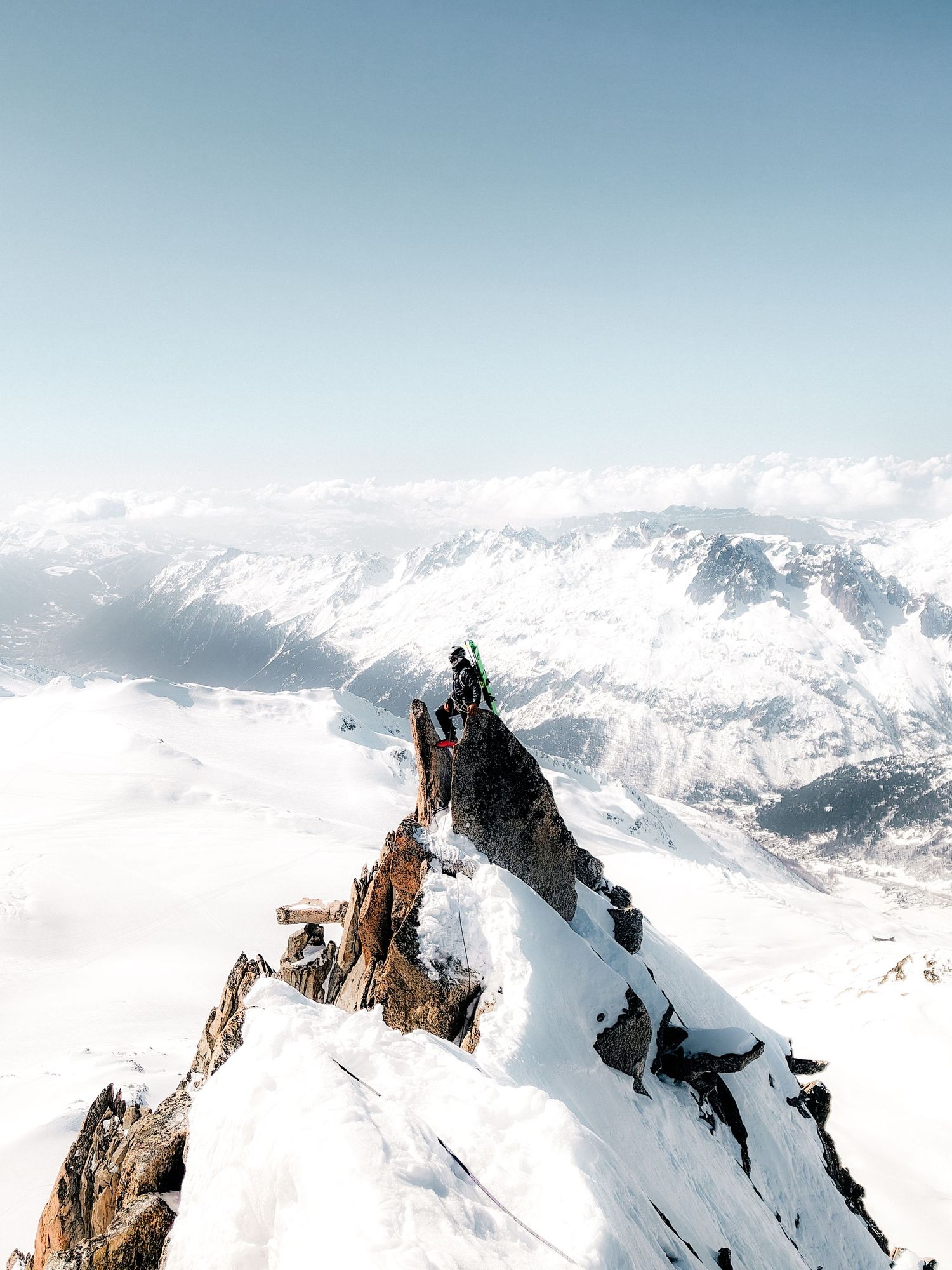 A climber stands atop a snow-covered rocky mountain peak with vast alpine landscape and misty mountains in the background.