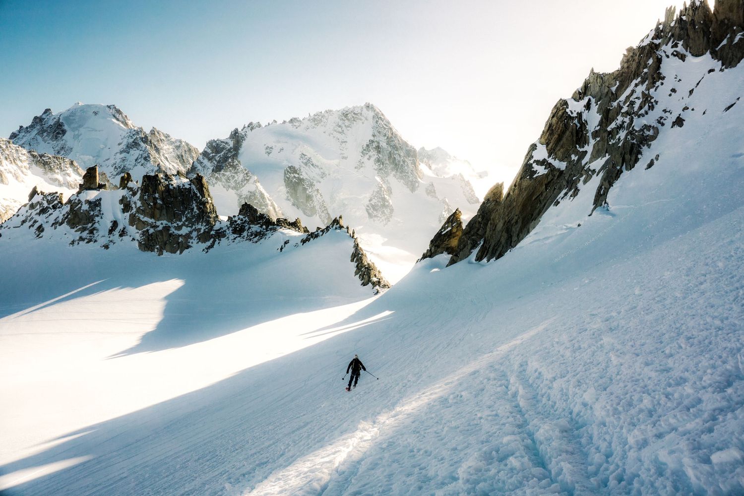 A lone skier descends a vast snowy alpine slope with dramatic sunlit mountain peaks in the background.