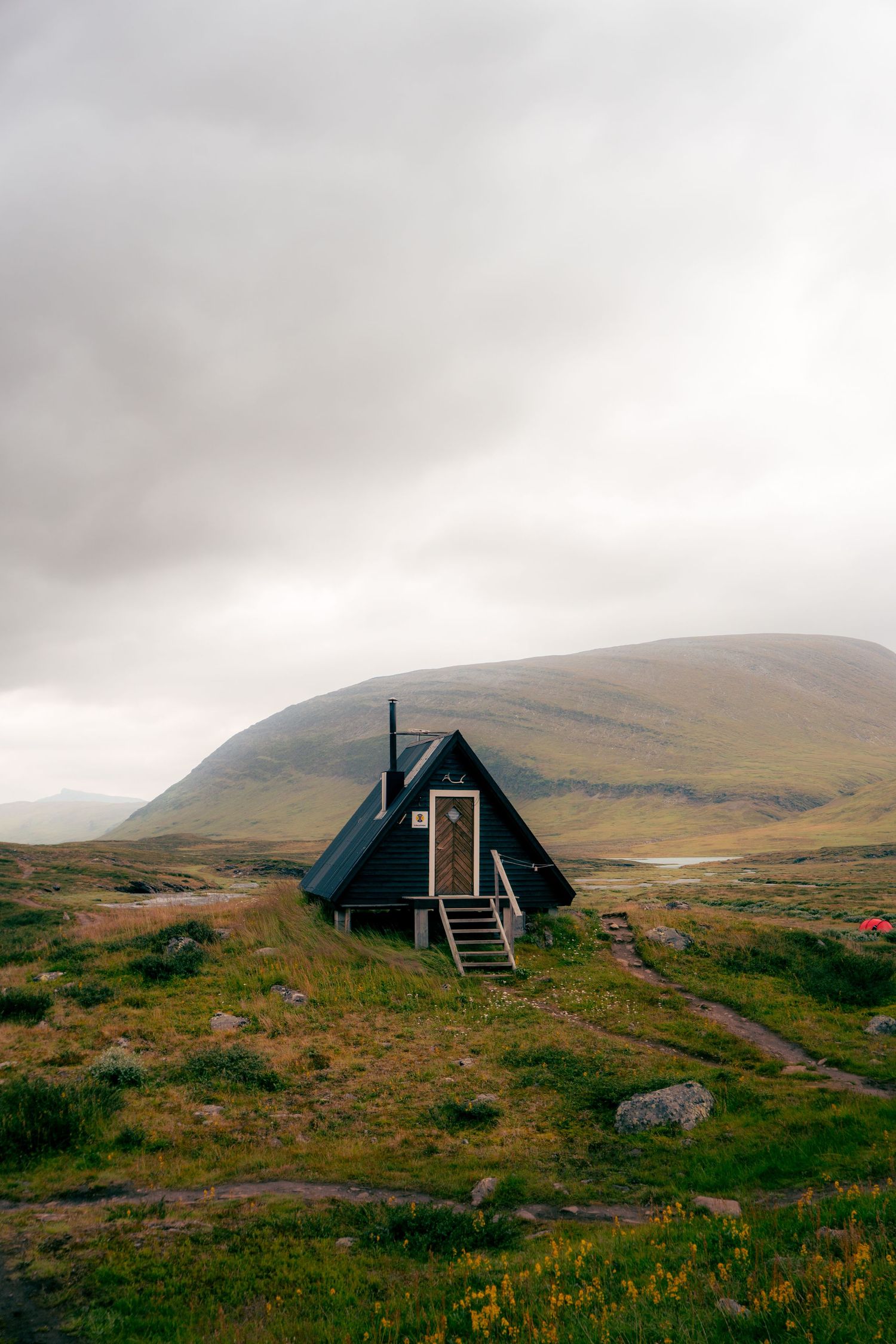 Black A-frame cabin with wooden door and steps on a green hillside with misty mountains behind.