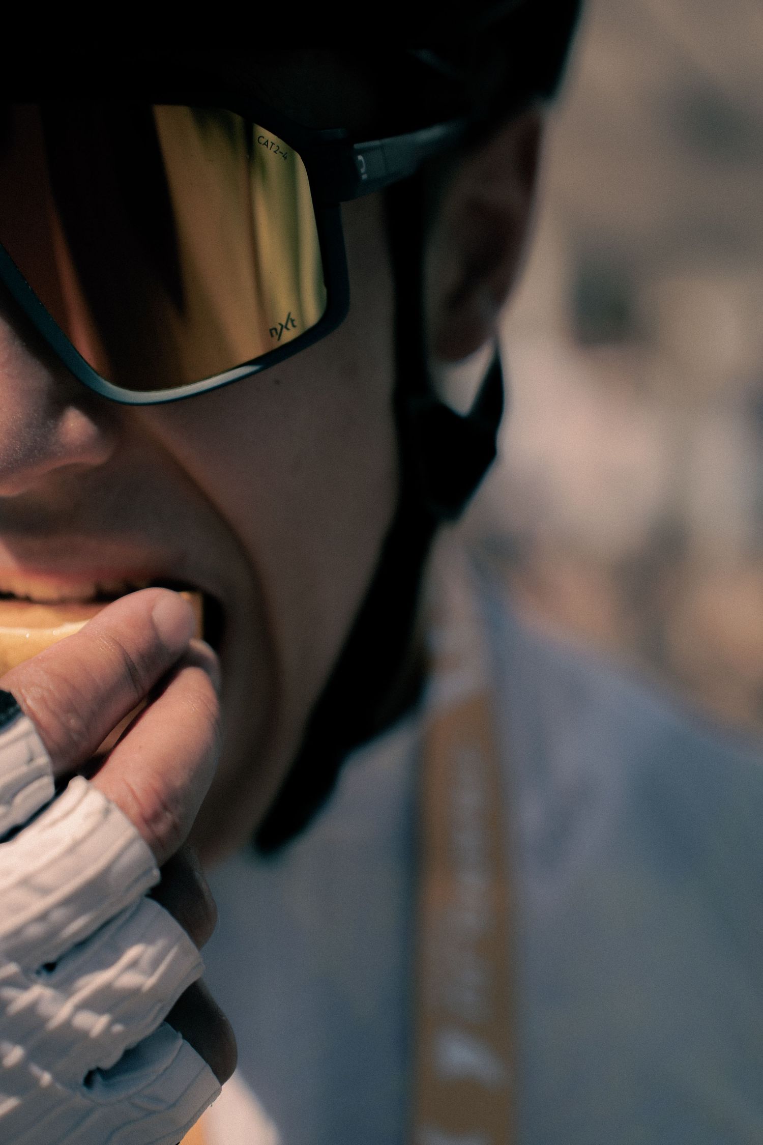 Cyclist wearing a black helmet and gold-mirrored sunglasses eating a snack with white cycling gloves.