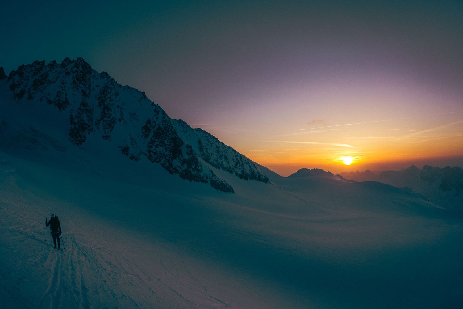 A lone skier ascends a snowy mountain slope at sunset with warm orange hues glowing on the horizon.