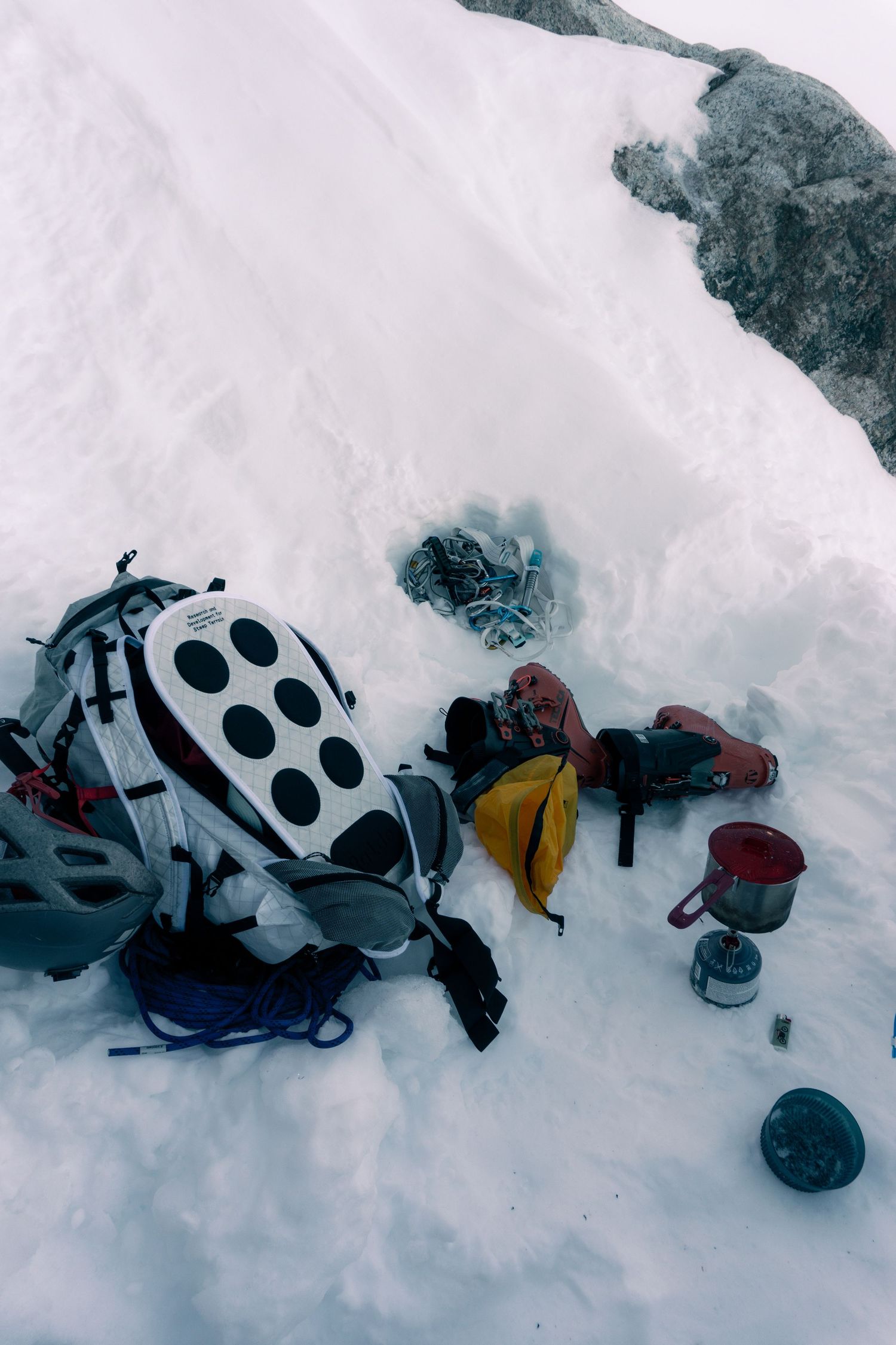 Mountain climbing gear including backpack, sleeping bag, and camp stove resting on snowy alpine terrain.