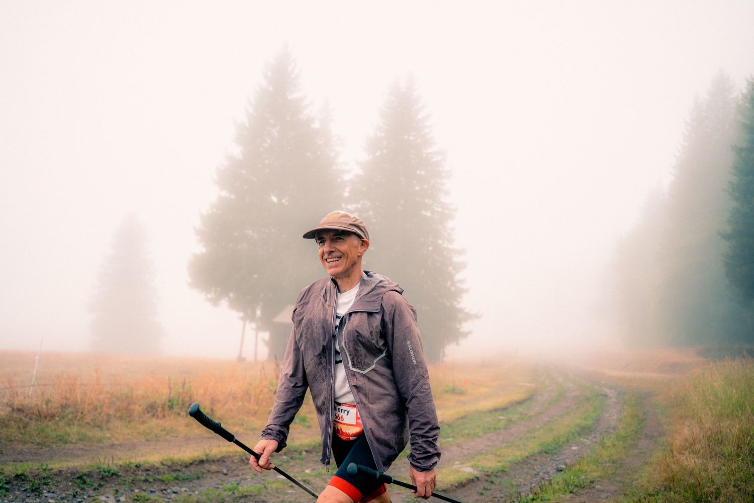 Smiling trail runner holds trekking poles on foggy mountain meadow trail.