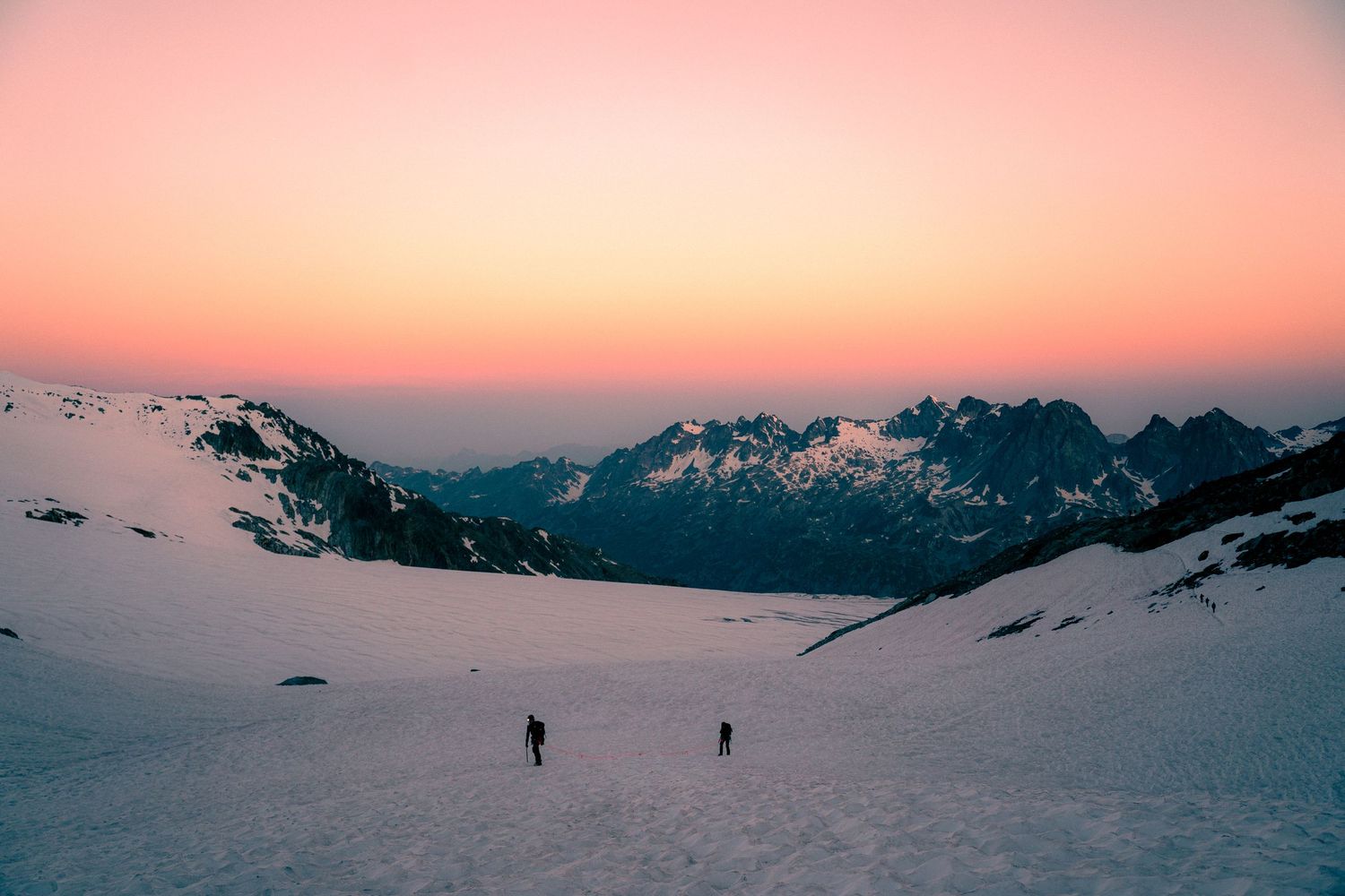 Two hikers traverse vast snow-covered glacier bowl beneath vibrant pink and orange sunset mountain sky.