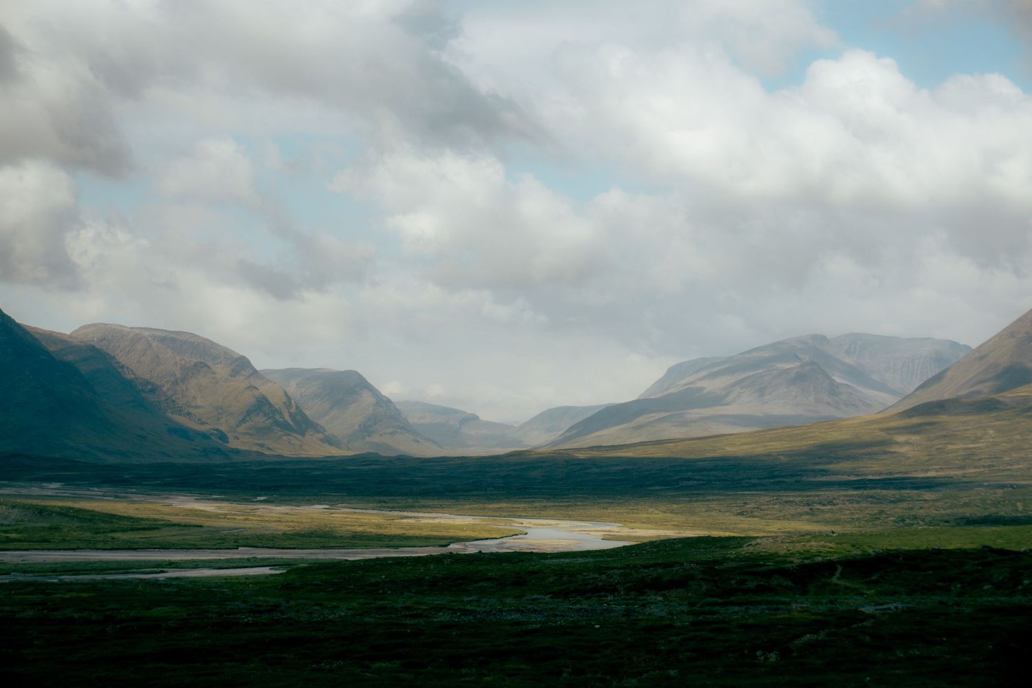Dramatic highland valley landscape with green meadows, a winding river, and misty mountains under cloudy skies.