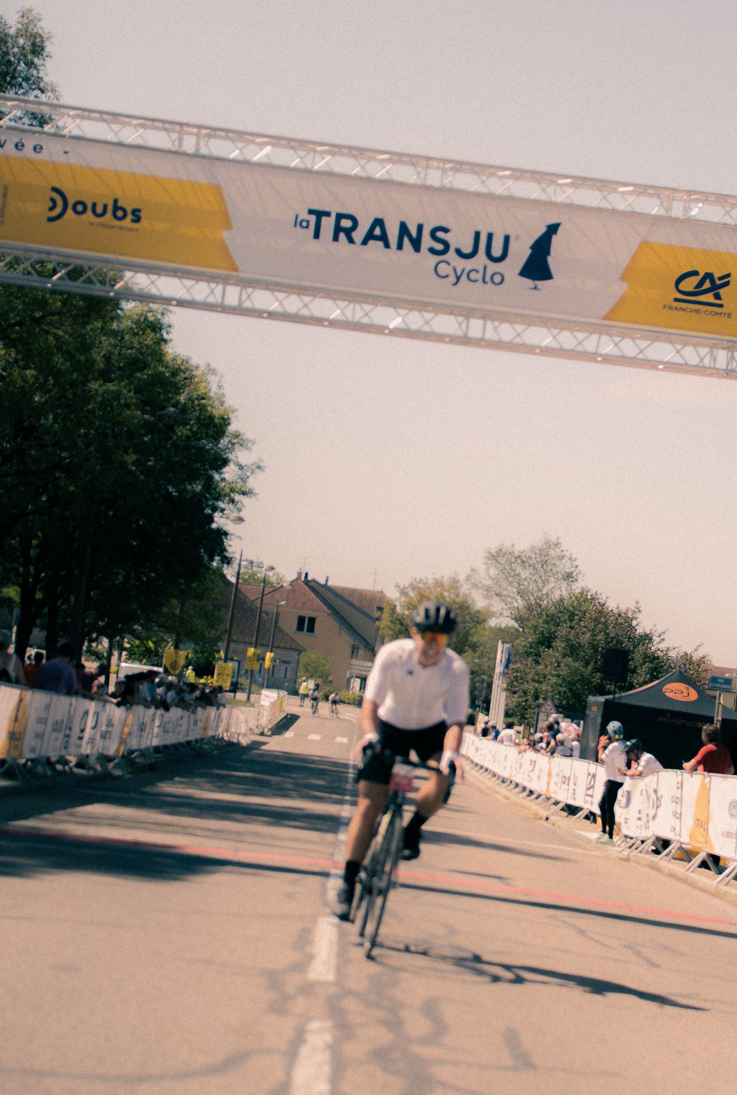 Cyclist crossing the finish line at La TransJu Cyclo race in Doubs, France, under a yellow and white banner.